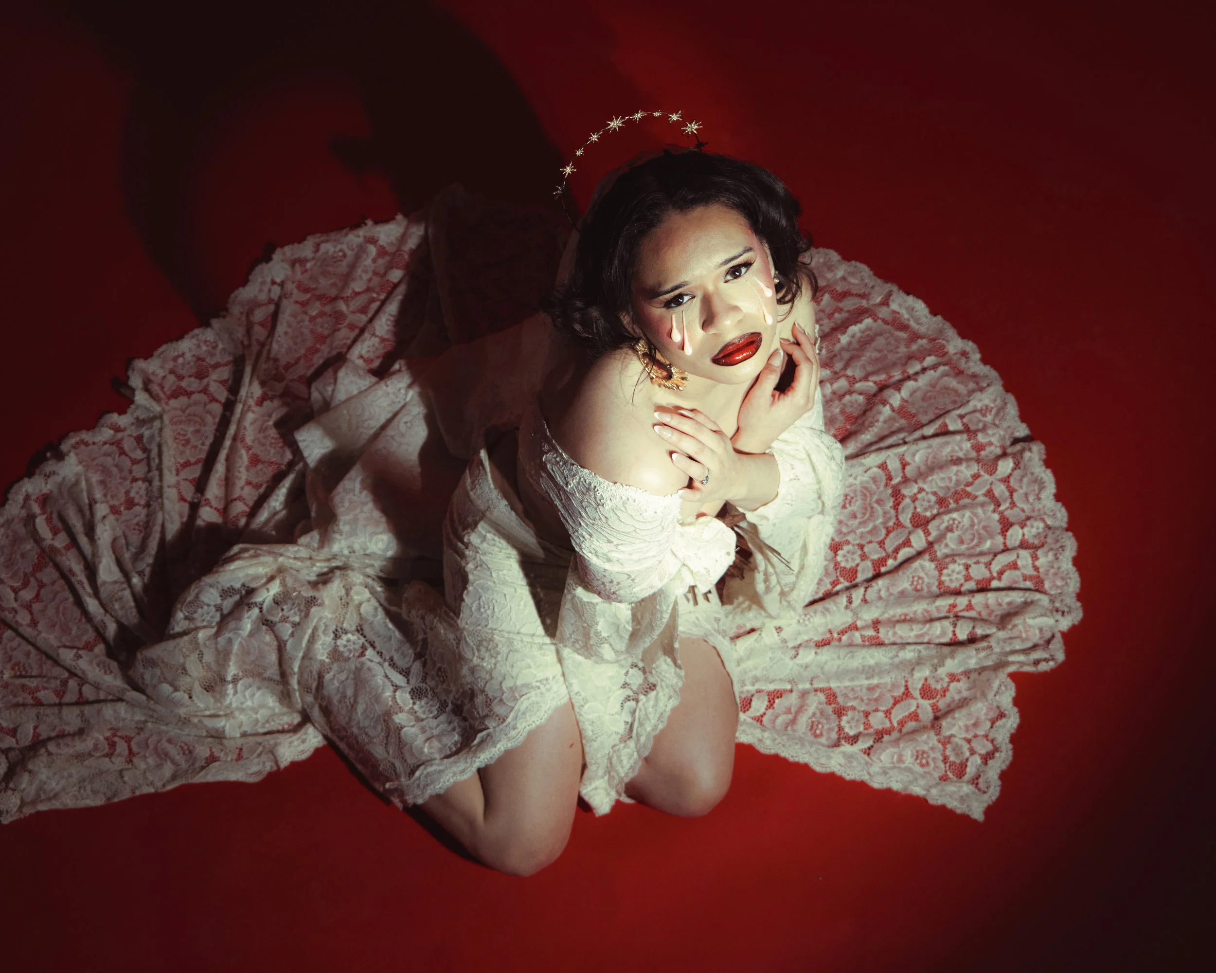 A woman with dark hair and makeup, Local Dallas, Texas, DFW Non-traditional Editorial Creative Portraits Cover Art Photographer. @roseypears .wearing a crown and a lacy white dress, is on her knees on a red surface, looking up at the camera with tear