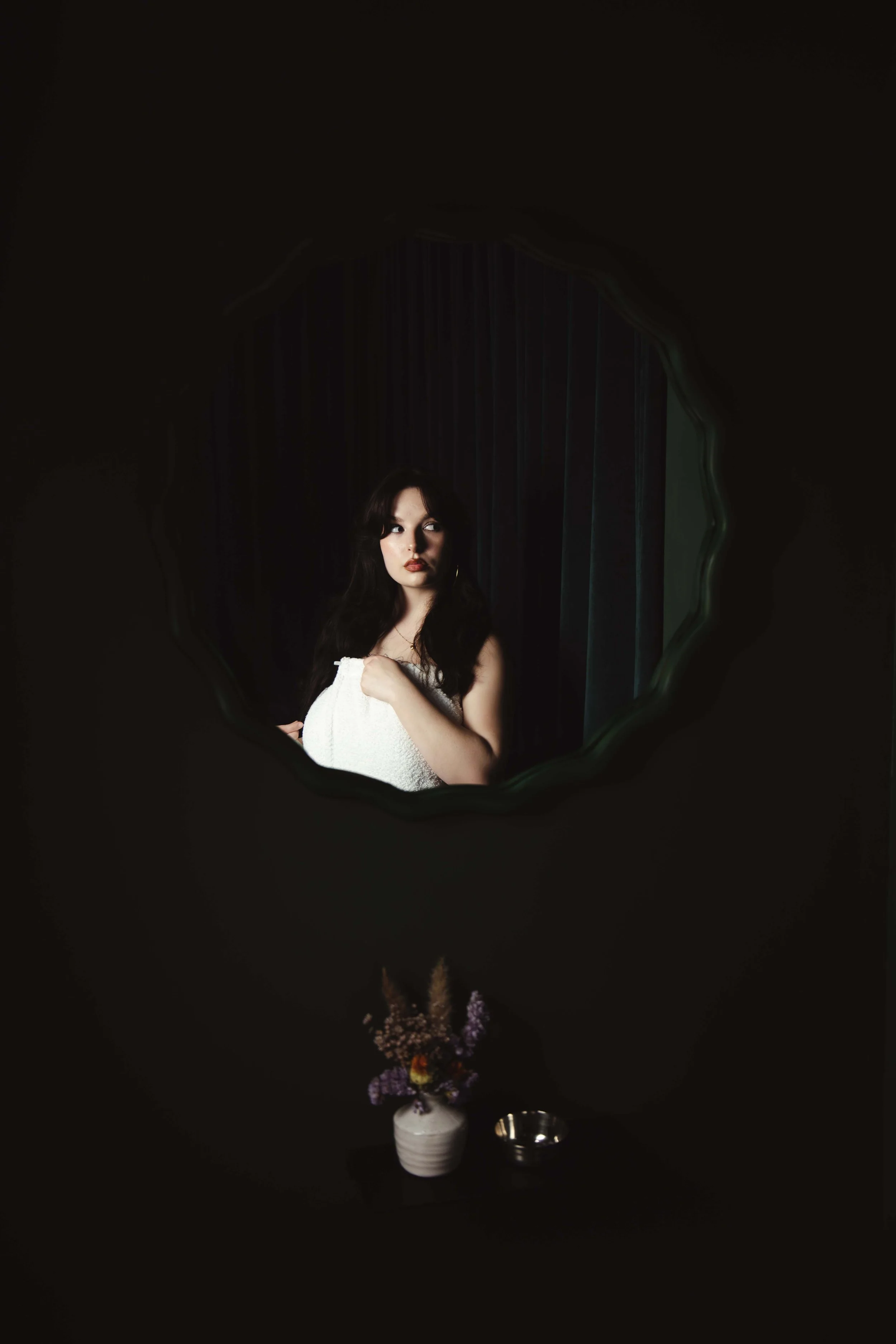 A woman with dark hair and red lipstick looking into a mirror framed with a dark, wavy border. In front of her is a table with a small vase of purple and orange flowers and a silver cup.