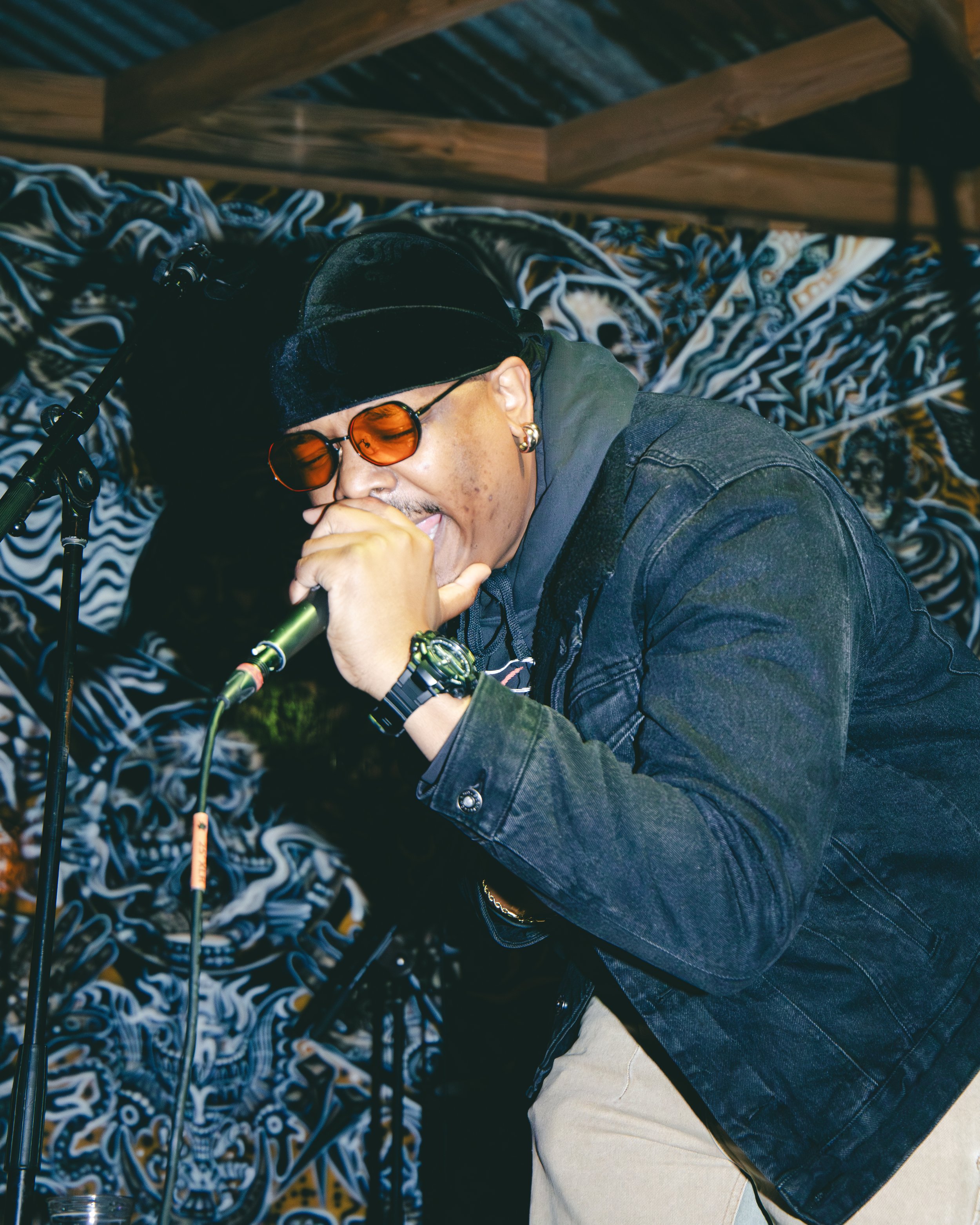 A man wearing orange-tinted sunglasses, a black cap, a dark blue jacket, and beige pants, sitting with his head tilted backward and mouth open, possibly singing or shouting, in front of a patterned background with blue, black, gold, and white designs