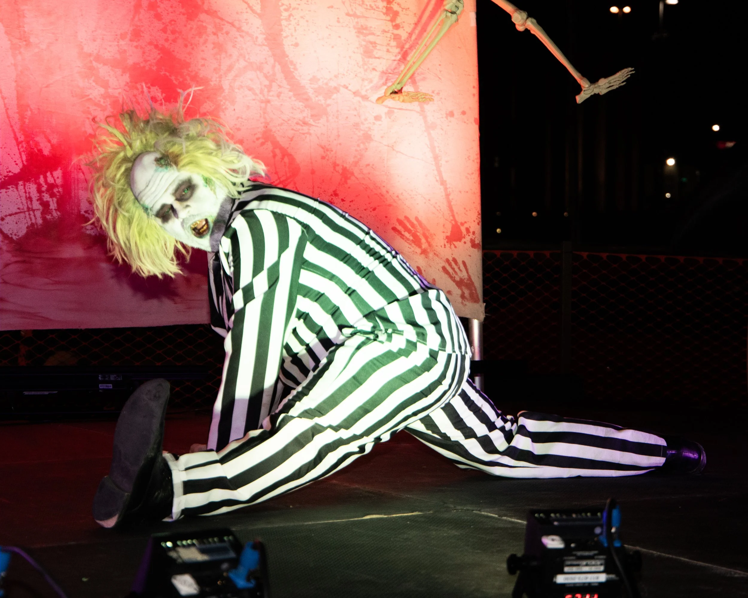 A performer with yellow hair and skeleton makeup, dressed in black and white striped clothing, posing on stage with a red horror-themed background. The stage has a black floor, and there are some electronic devices in the foreground.