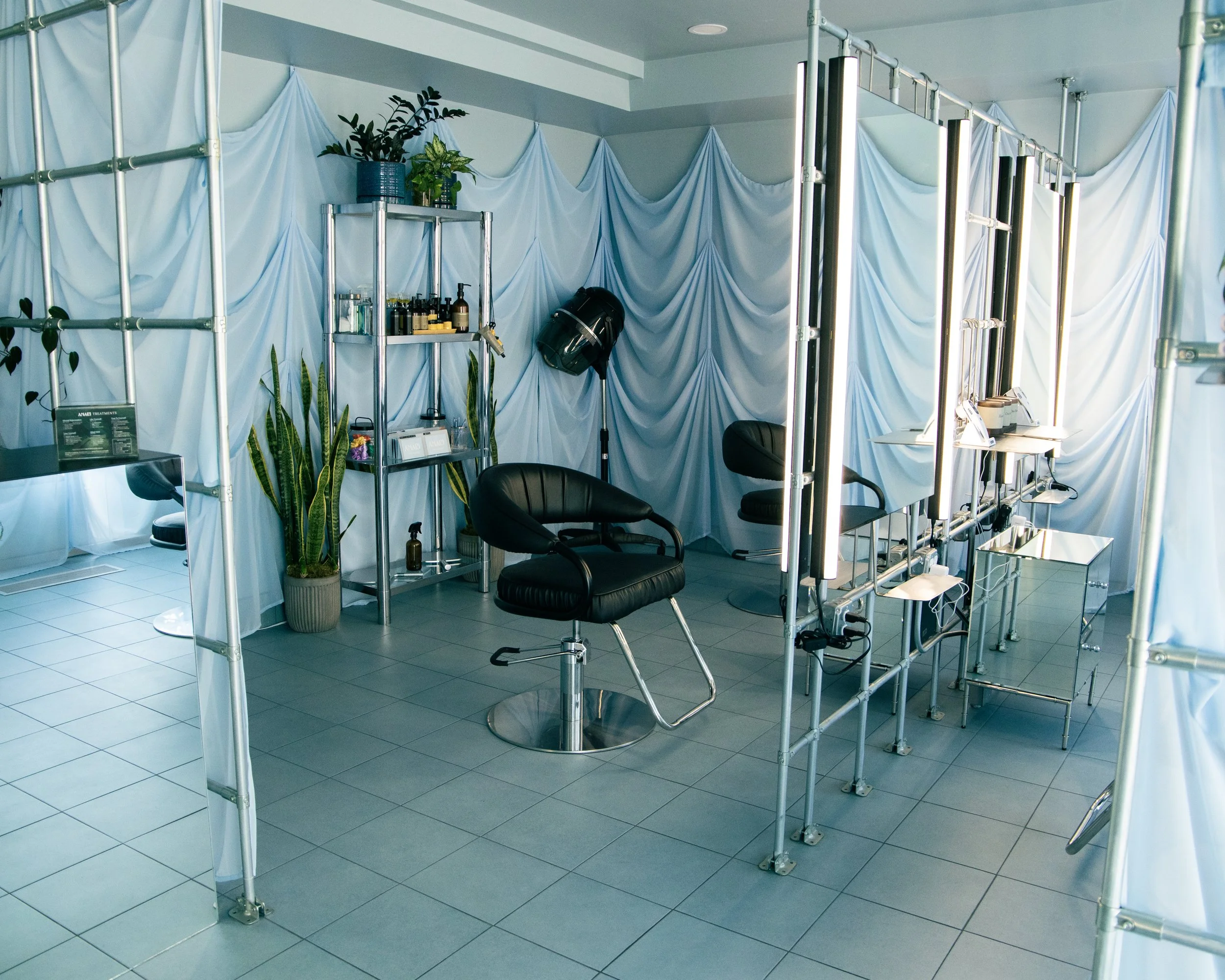 Indoor space with a white draped backdrop, a black barber chair, plants, and a metal shelving unit with bottles, set up for a beauty or hair salon.