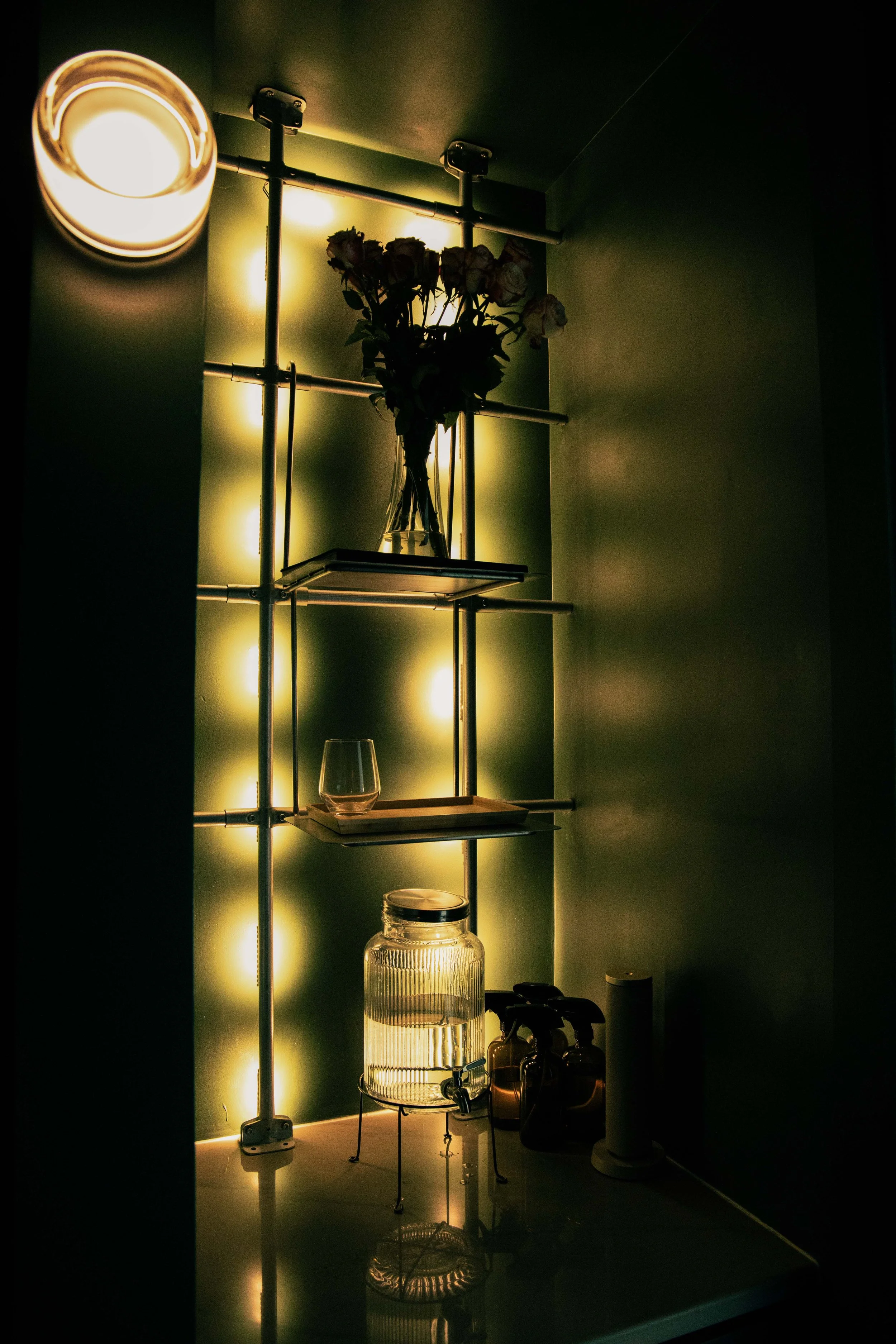 Dimly lit room with a metal shelf containing a bouquet of roses in a vase, a candle, and cleaning supplies on a counter. The back wall has yellow-green lighting creating a warm glow.