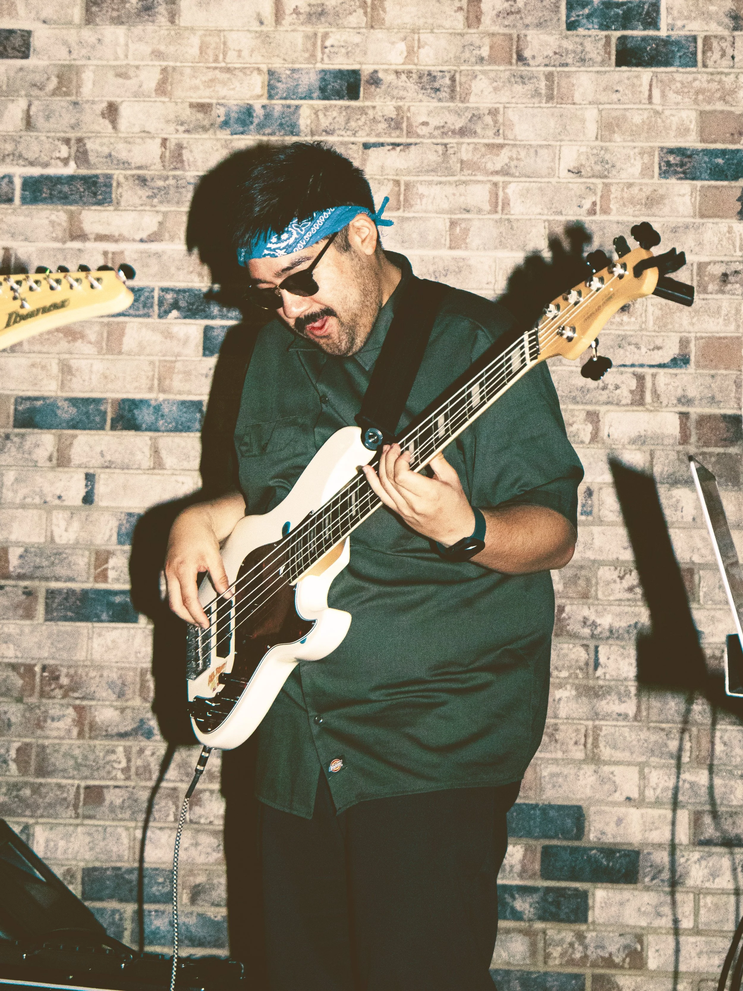 A man playing an electric guitar against a brick wall, wearing a blue bandana, sunglasses, and a dark shirt.