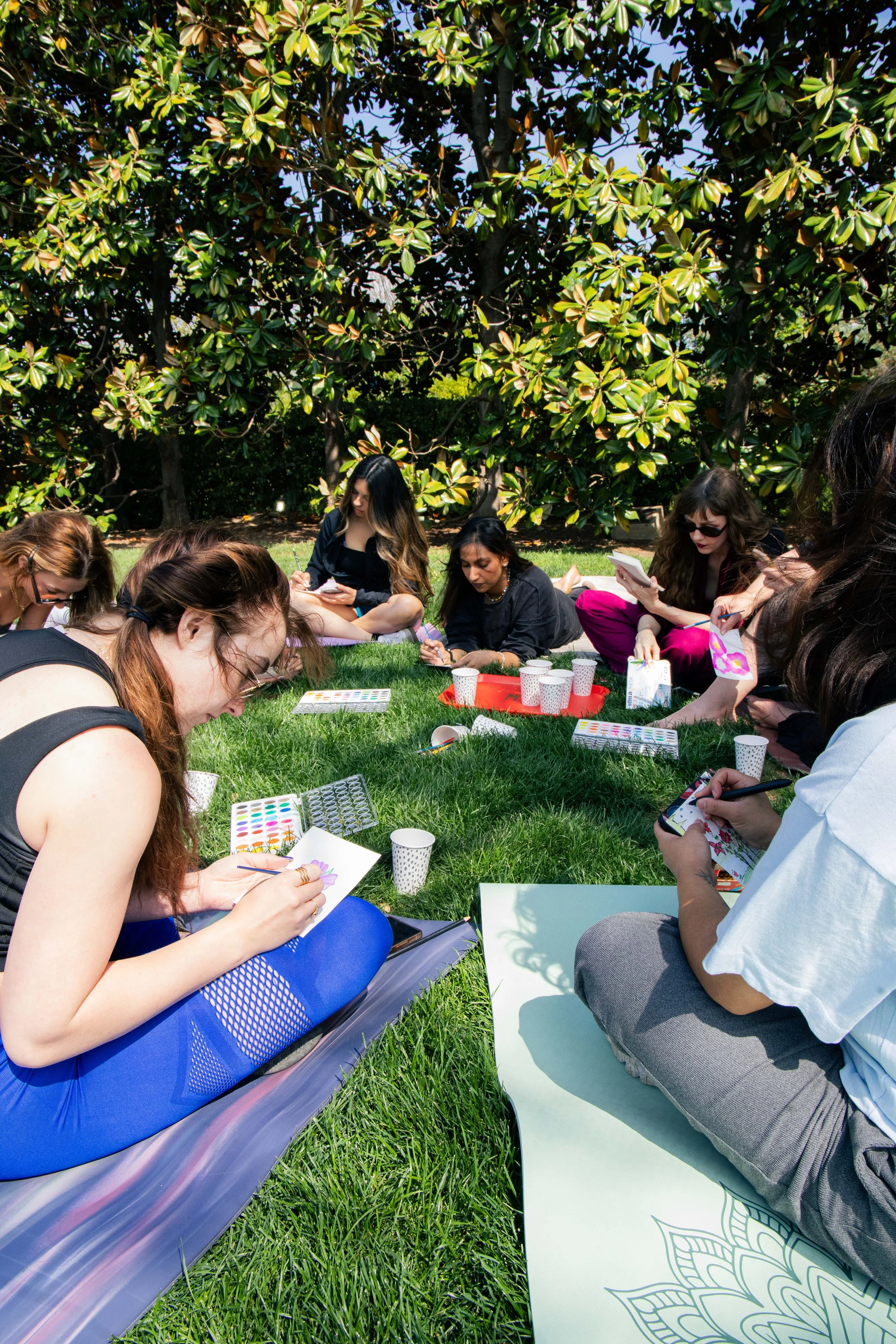 Group of women sitting outdoors on grass, engaging in arts and crafts activities, with large trees in the background.