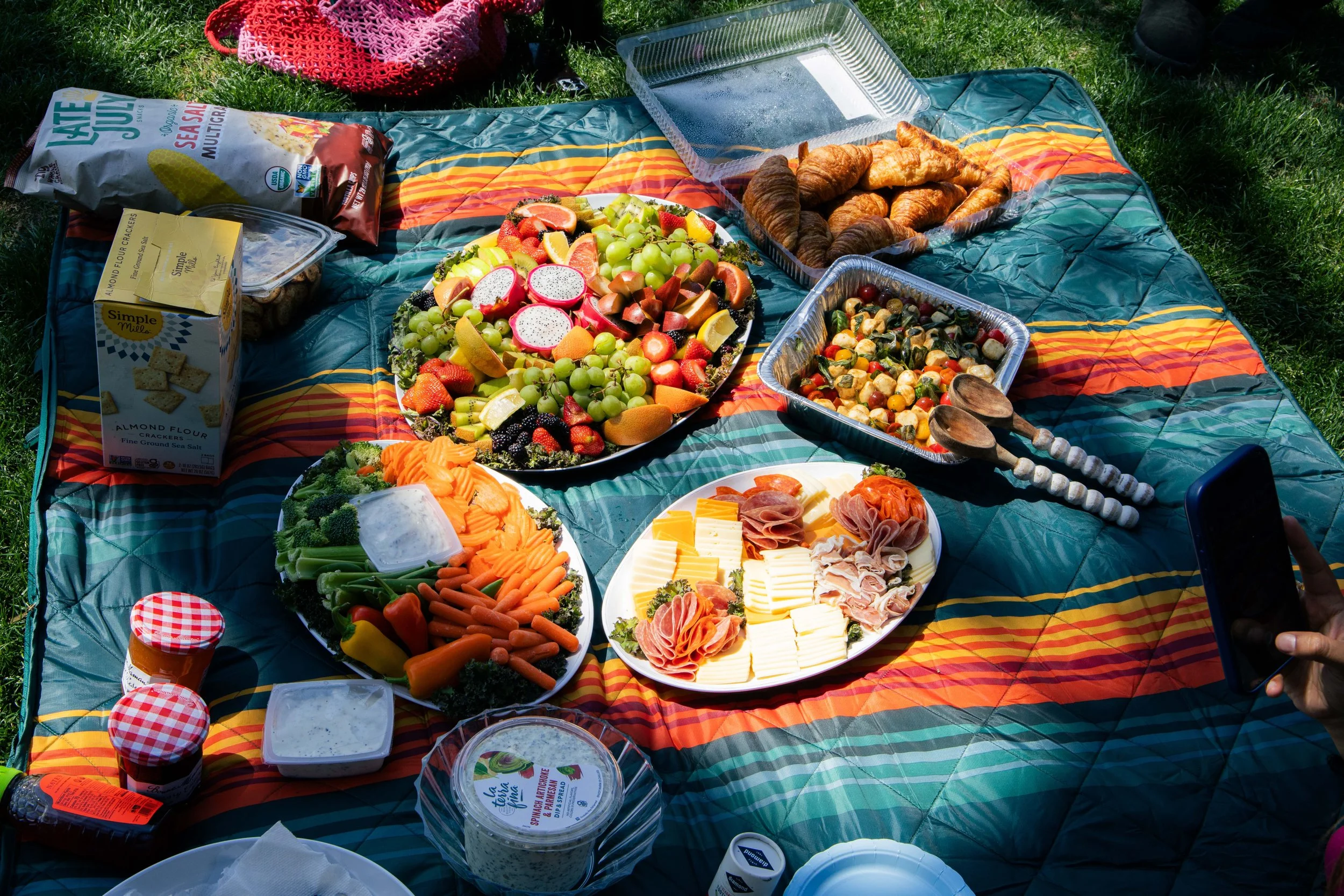 A picnic spread on a colorful blanket with various food items including a large fruit platter with grapes, dragon fruit, and berries, croissants, cheese and meats, vegetable tray with carrots, broccoli, and cherry tomatoes, and packaged snacks and de
