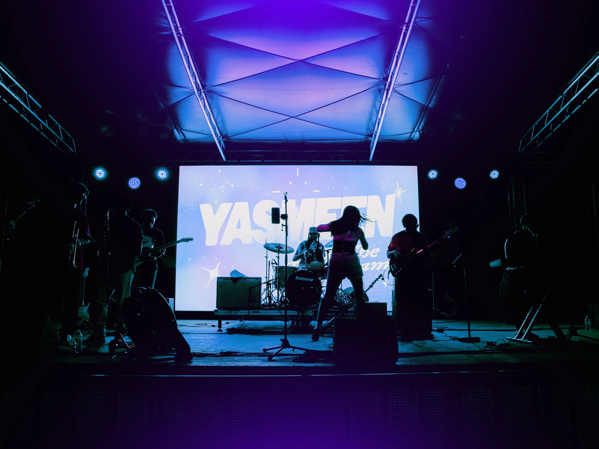 Stage with band performing under purple and blue lighting, large screen displaying text, and audience members silhouette in foreground. Local Dallas, Texas musicians.