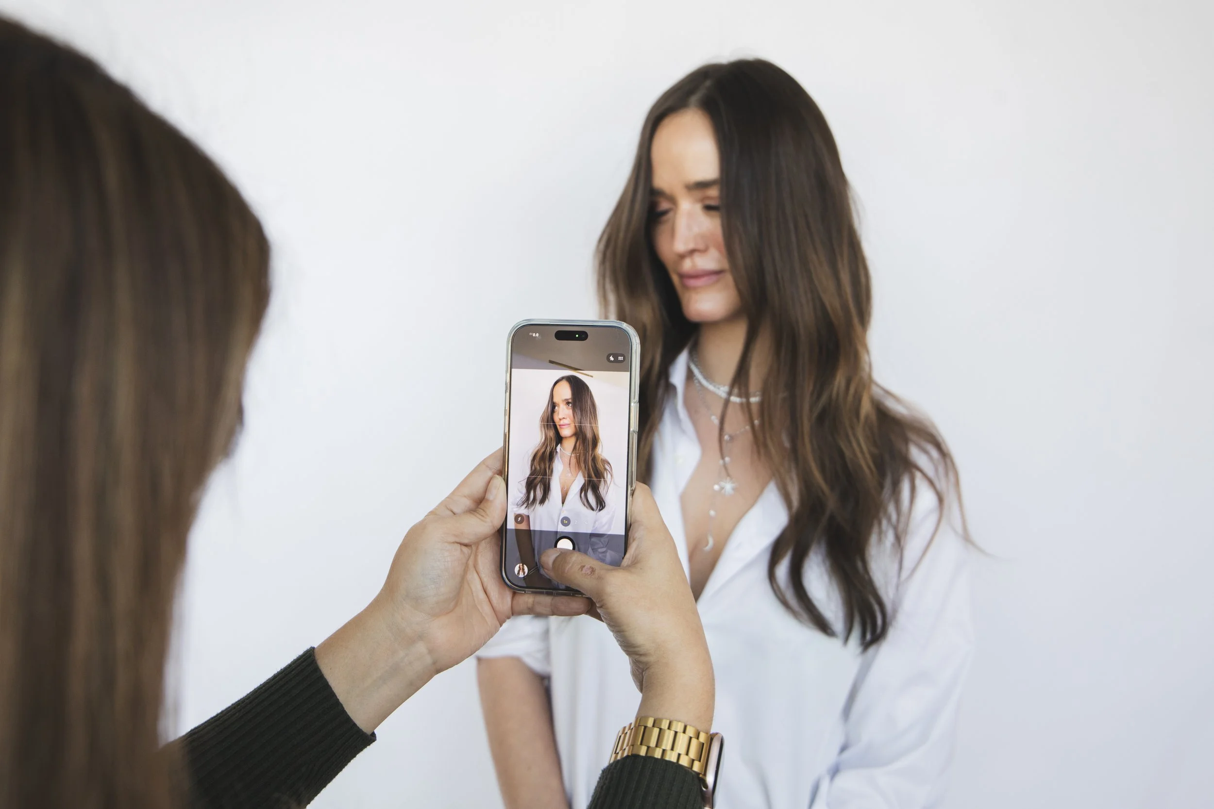 A woman with long brown hair and a white shirt poses for a photo while another person takes her picture with a smartphone.