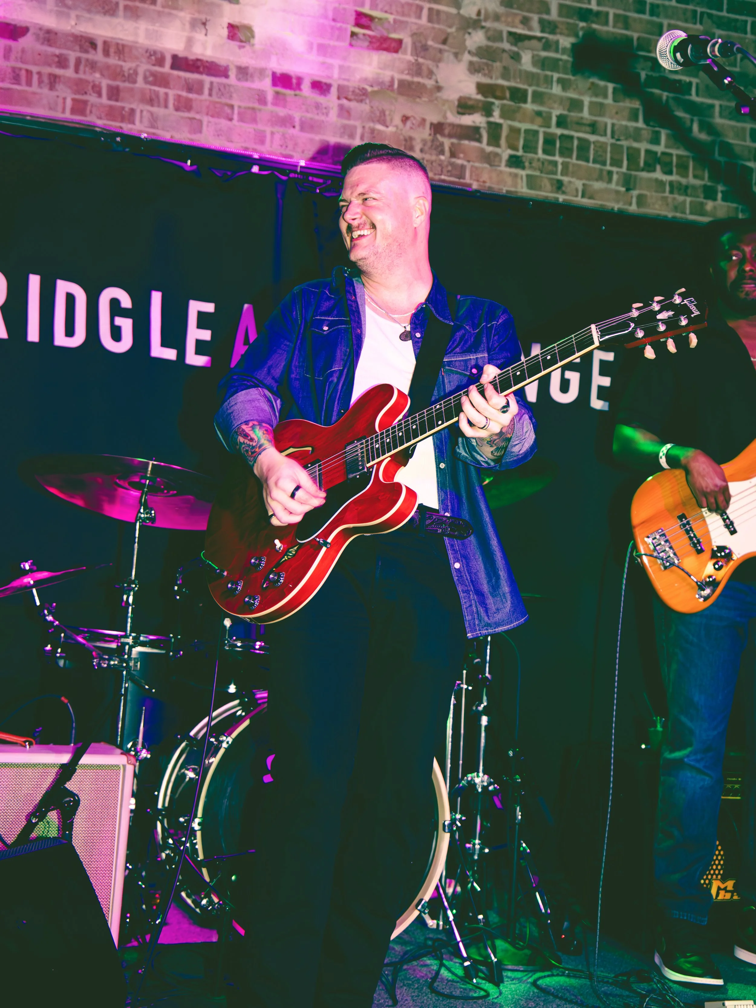 A man playing an electric guitar on stage with a brick wall behind him, smiling and enjoying the music. There are drum set components and another person with an acoustic guitar in the background.