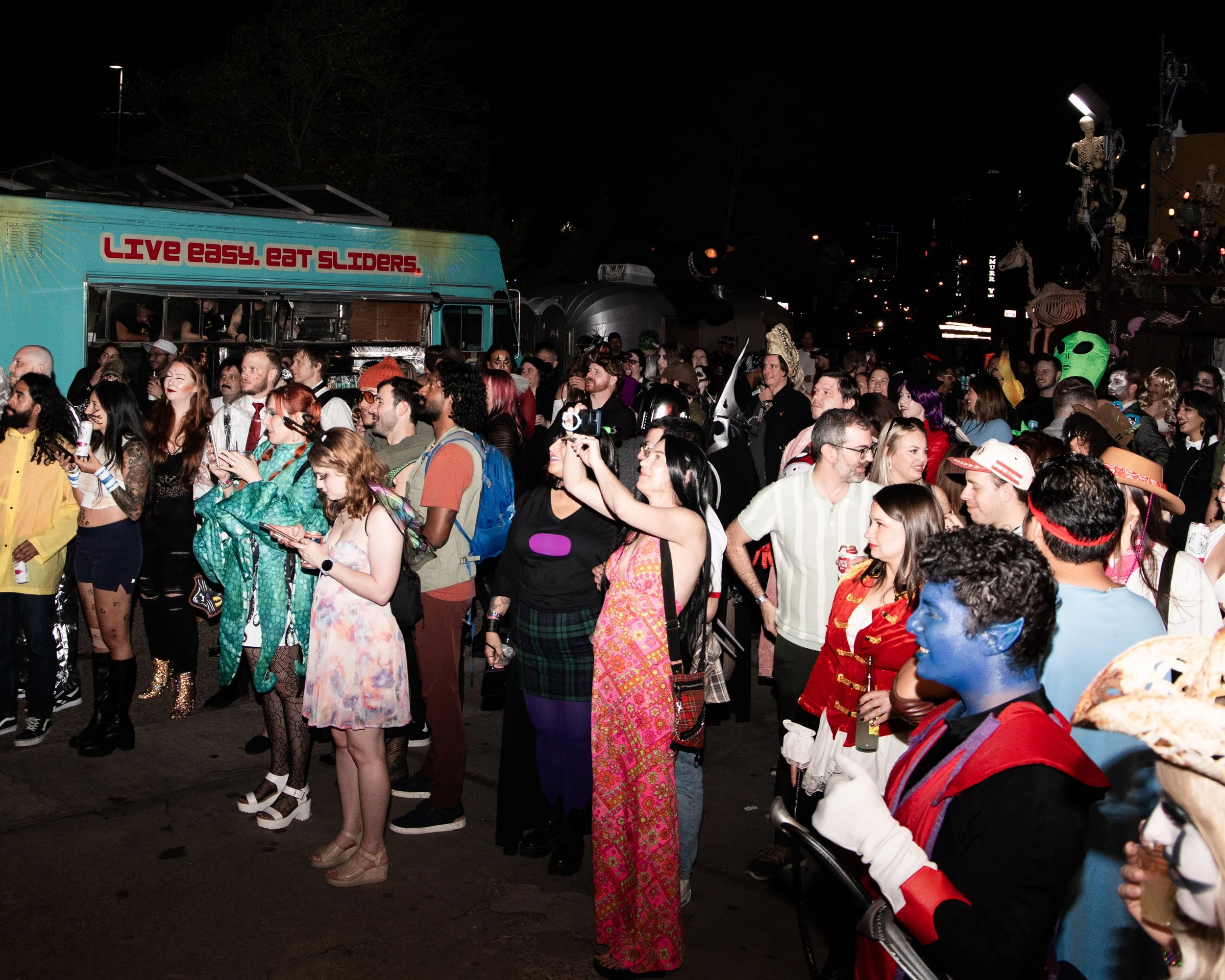 Crowd of people dressed in costumes at night, with some smiling and taking photos, in front of a food truck and Halloween decorations.