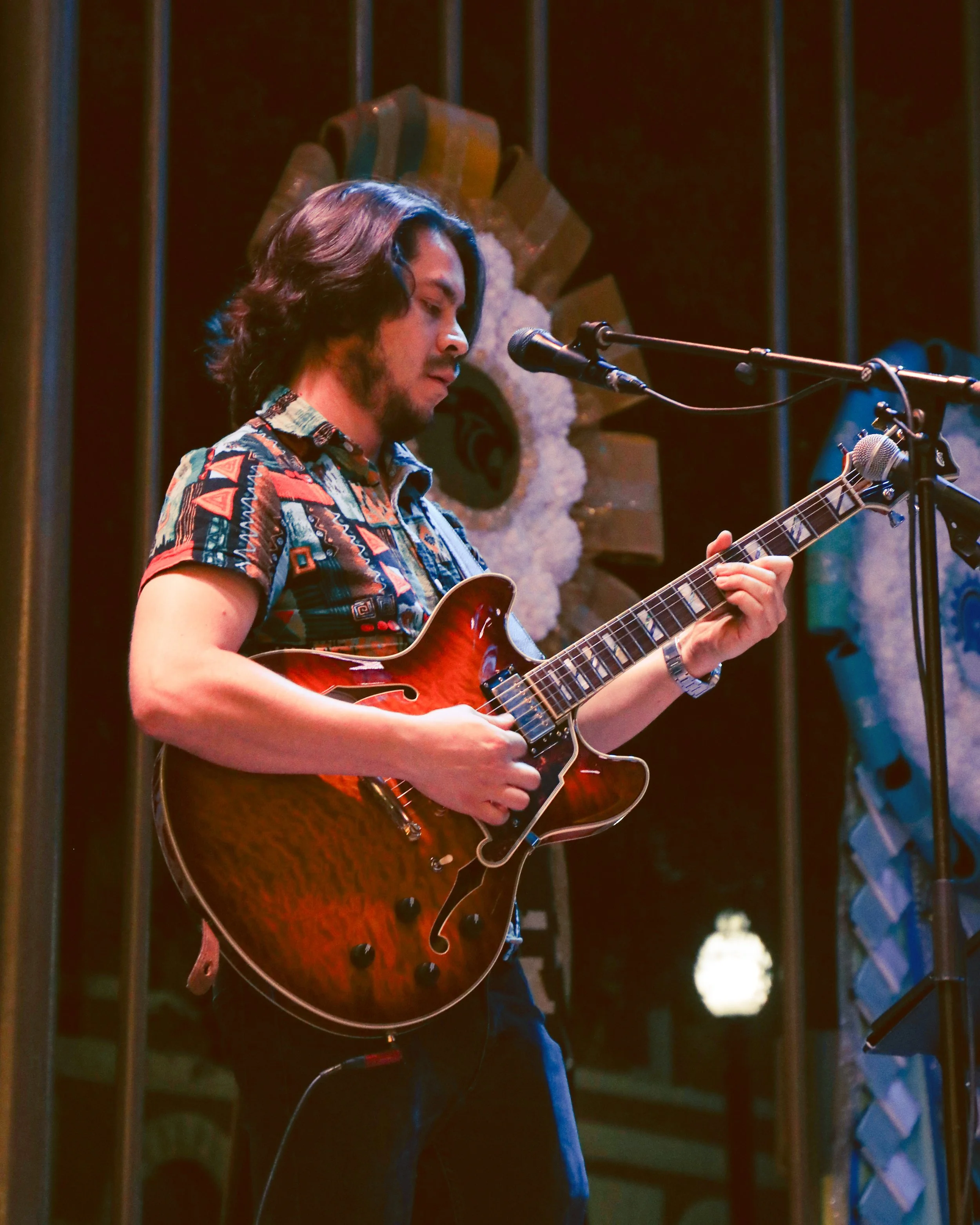 A man with long dark hair and beard playing an electric guitar on stage, with microphones set up in front of him. The background features wooden panels and a floral decoration.