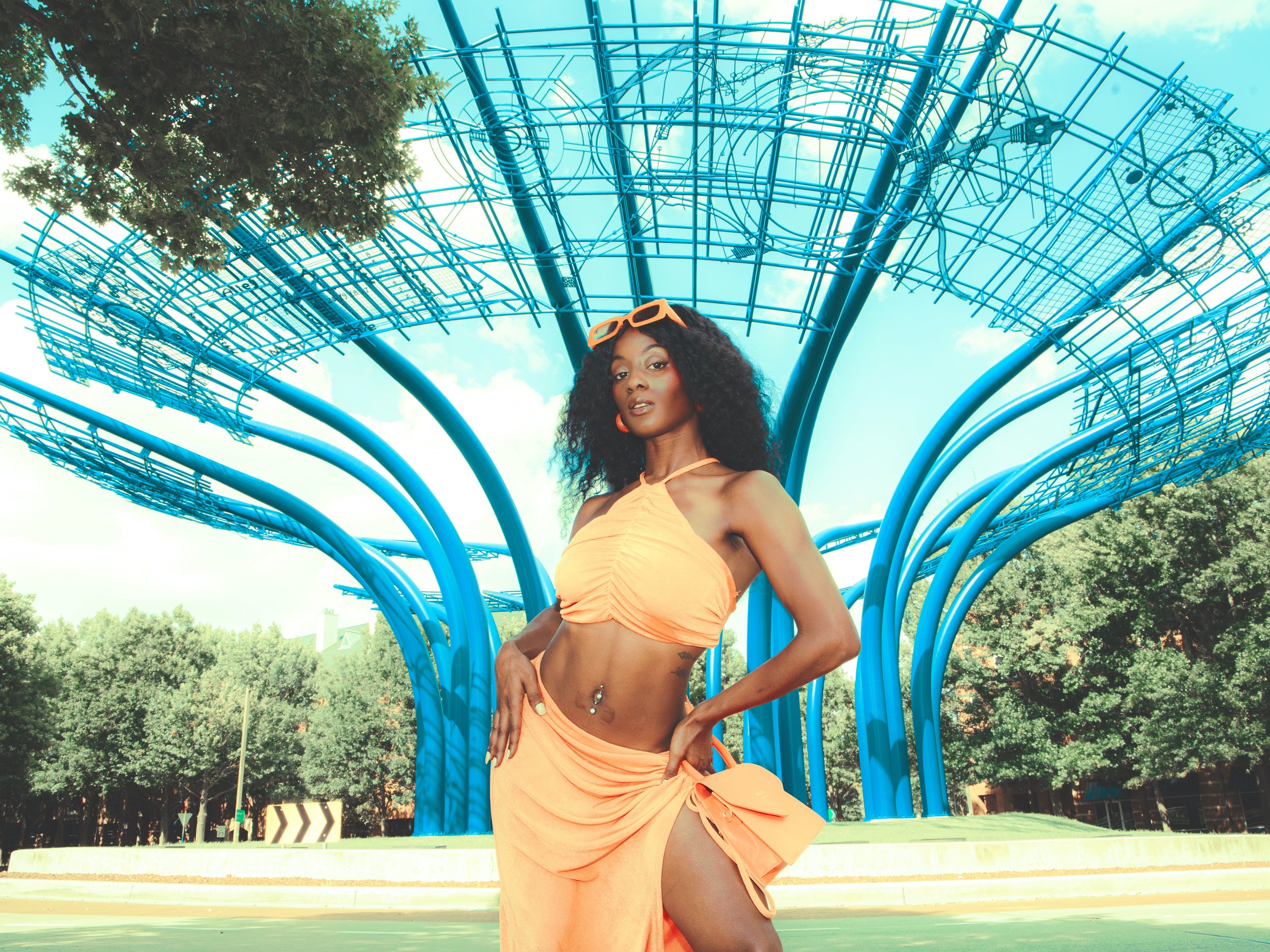 Local Dallas, Texas, DFW Non-traditional Fashion Editorial Creative Portraits Cover Art Photographer Photoshoot. Addison Circle. A woman with curly black hair and orange sunglasses stands outdoors in front of a blue metal sculpture resembling a tree 