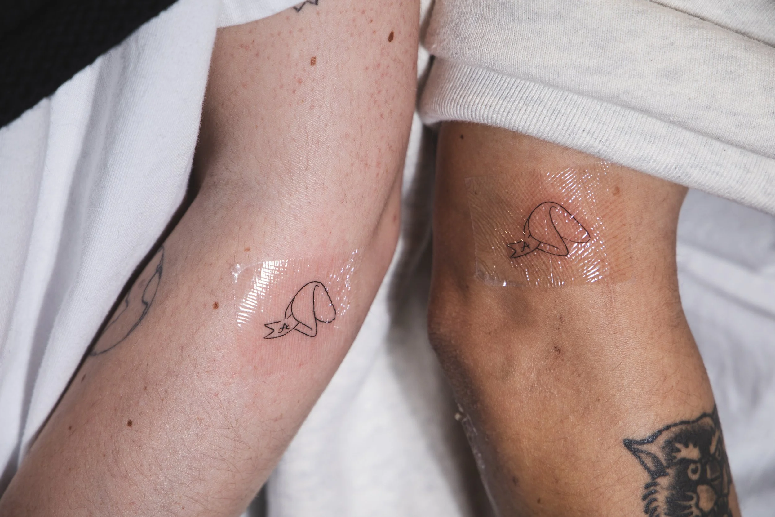 Close-up of two arms with tattoos, one with freckles and the other with a henna-style tattoo, both with small bandages and transparent tape on the skin.