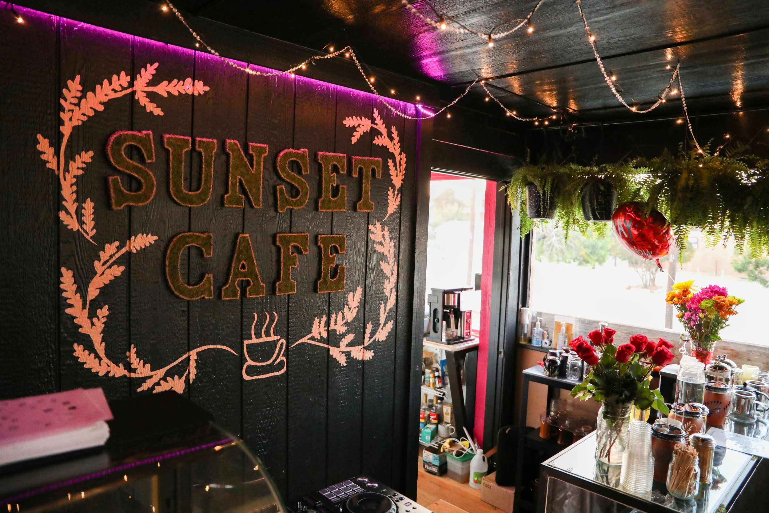 Interior view of a cafe with a large black wall decorated with pink lettering that reads "Sunset Cafe" surrounded by pink leaf illustrations. The ceiling is adorned with string lights and hanging greenery. There are shelves with jars, cups, and plant