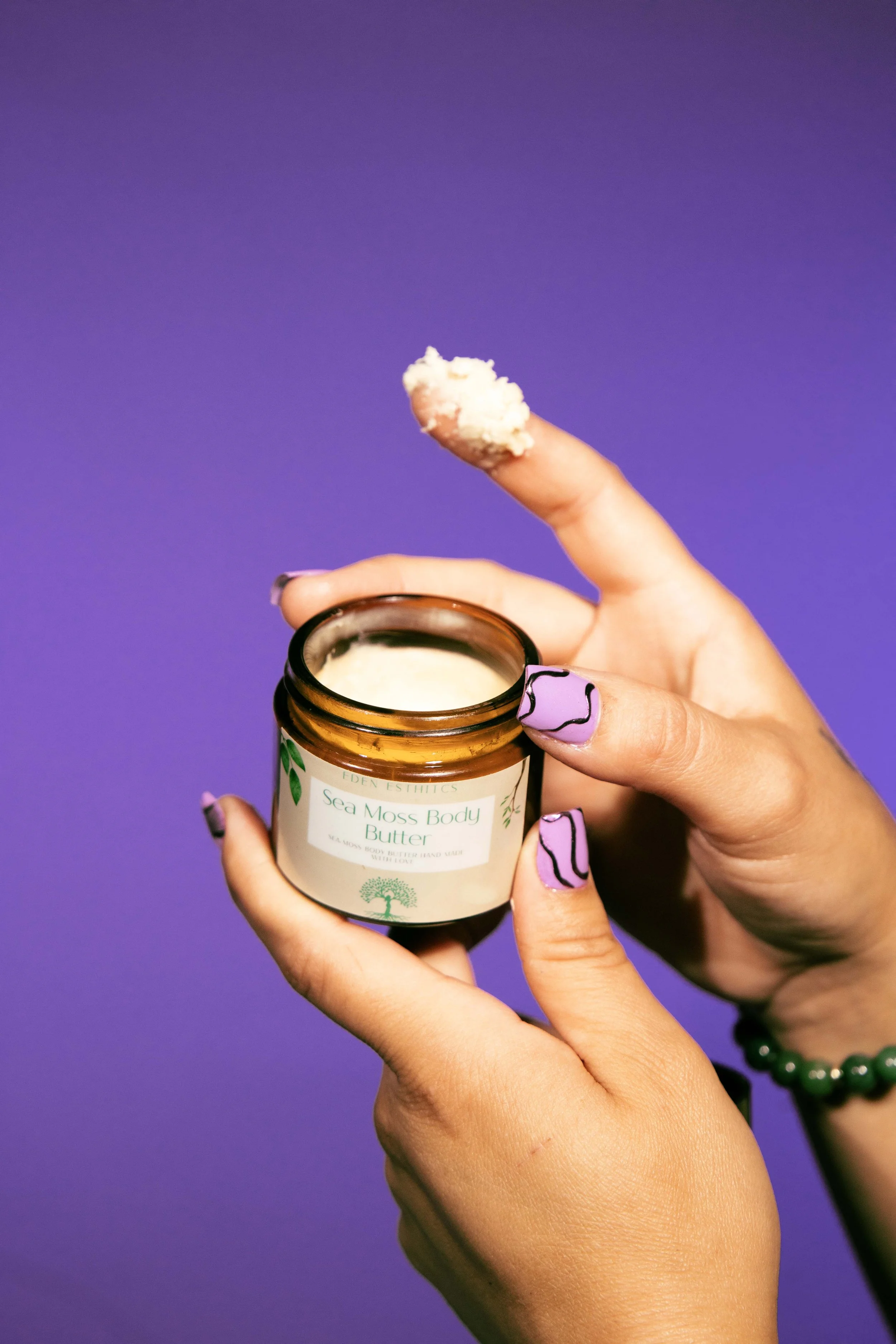 Person holding an open jar of Eden Esthtics Sea Moss Body Butter against a purple background