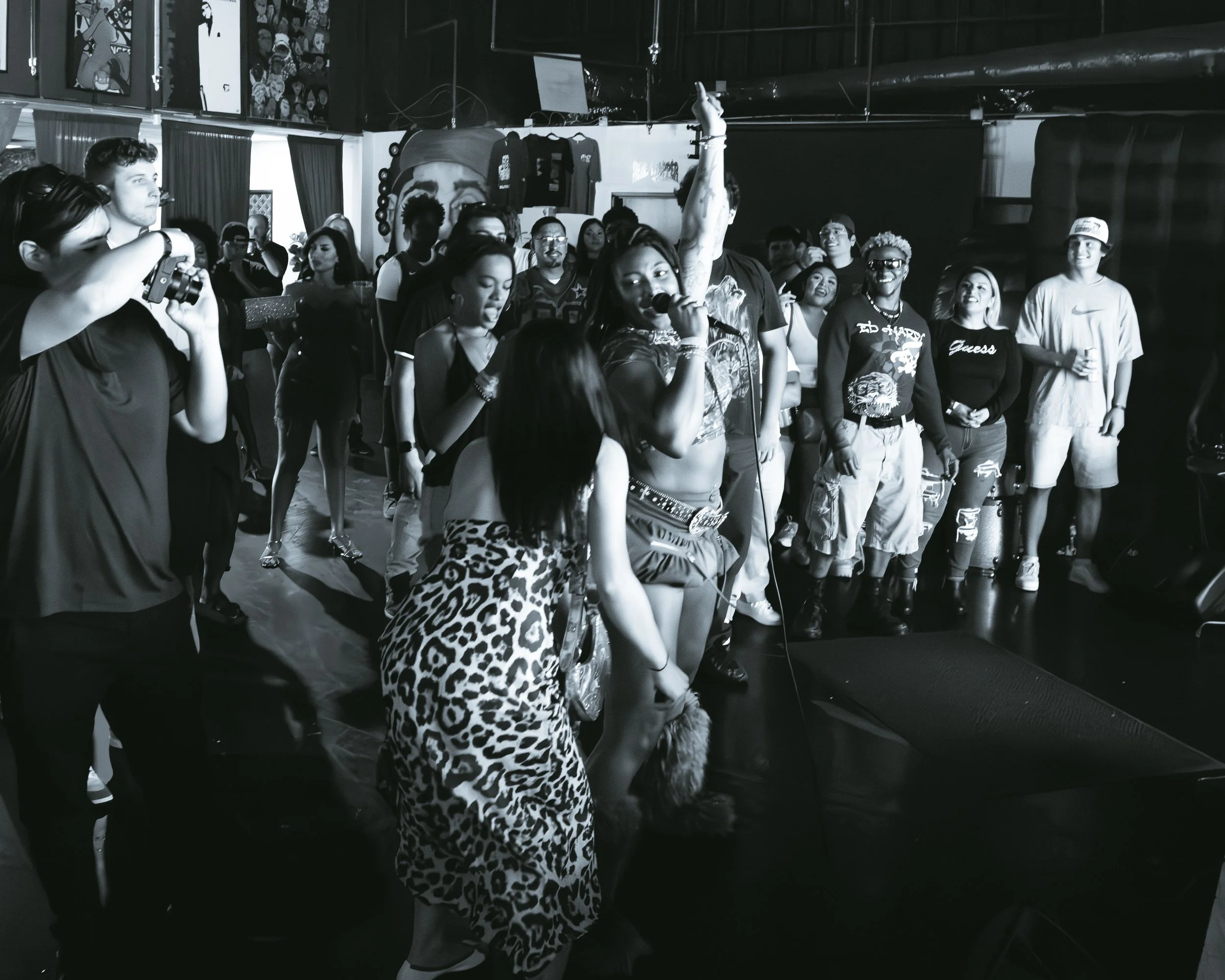 A group of diverse people enjoying a lively concert or event, with a woman singing into a microphone, others dancing, and some taking photos, in an indoor venue with wall art and dark lighting.