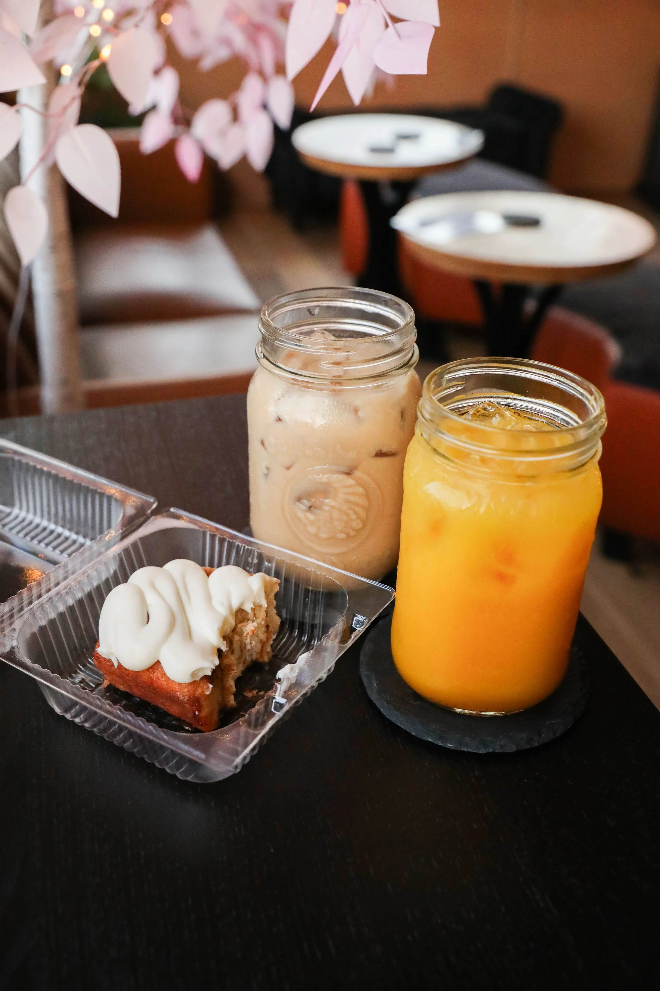 Sweet dessert cake with frosting, two mason jars with iced drinks, and a tray of food on a black table in a cozy cafe setting with pink flower decoration.