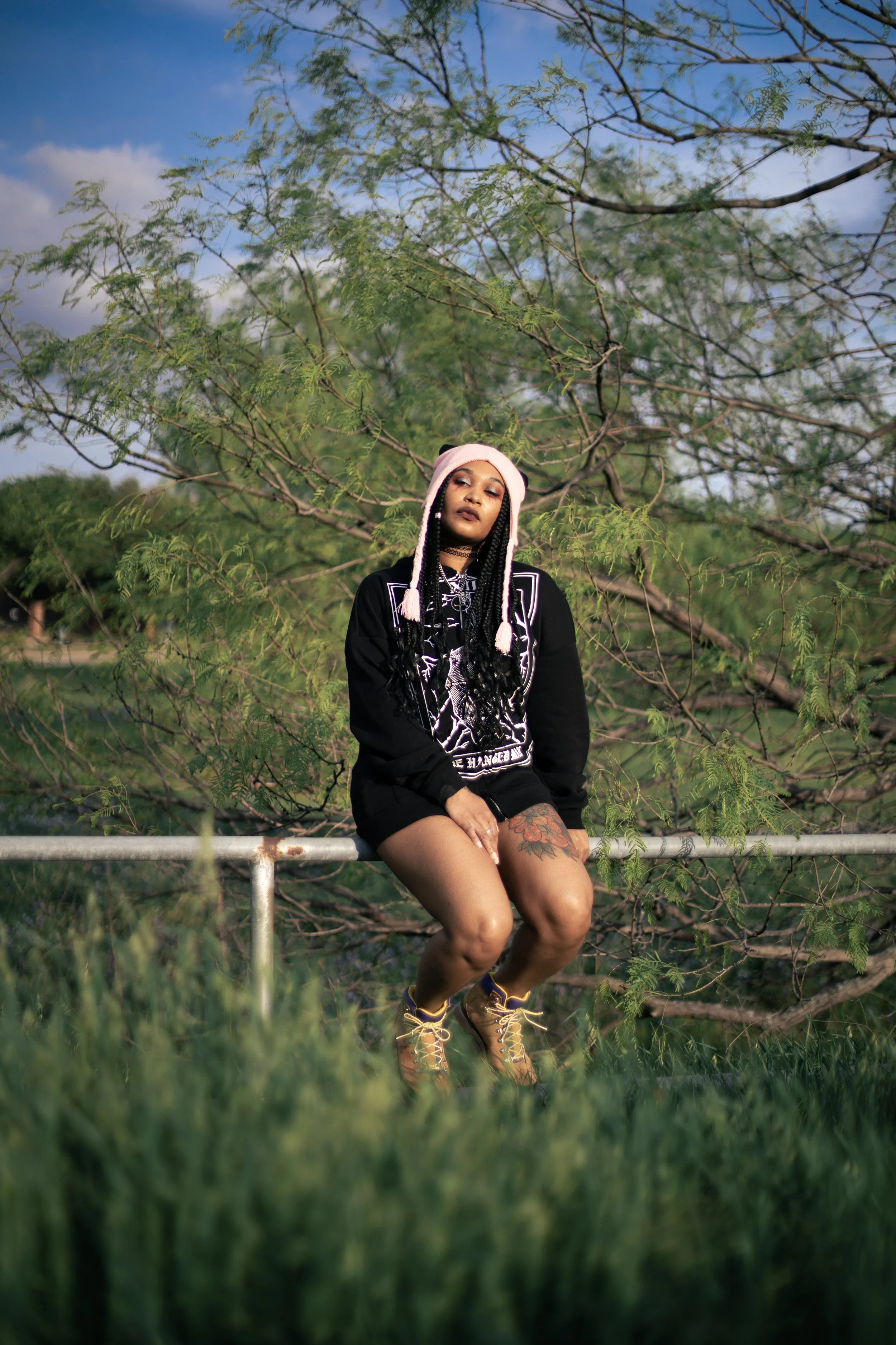 Local Dallas, Texas, DFW Non-traditional Fashion Editorial Creative Portraits Cover Art Photographer Photoshoot. musician igimeji .A woman with braided hair, wearing a pink hood and black hoodie, sitting on a metal railing in front of green foliage a