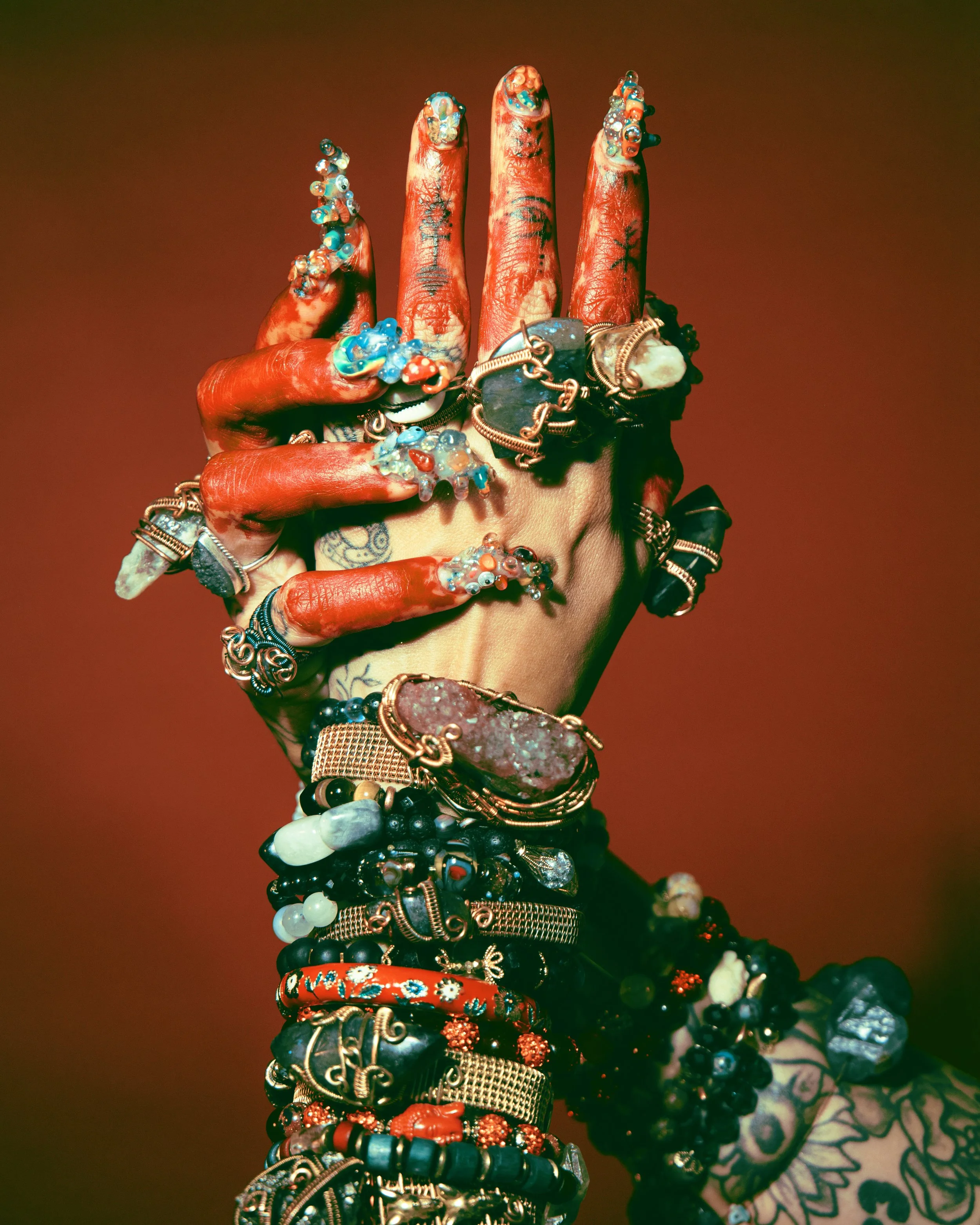 A hand with various colorful jewelry, rings, bracelets, and tattoos, posed against a warm brown background.