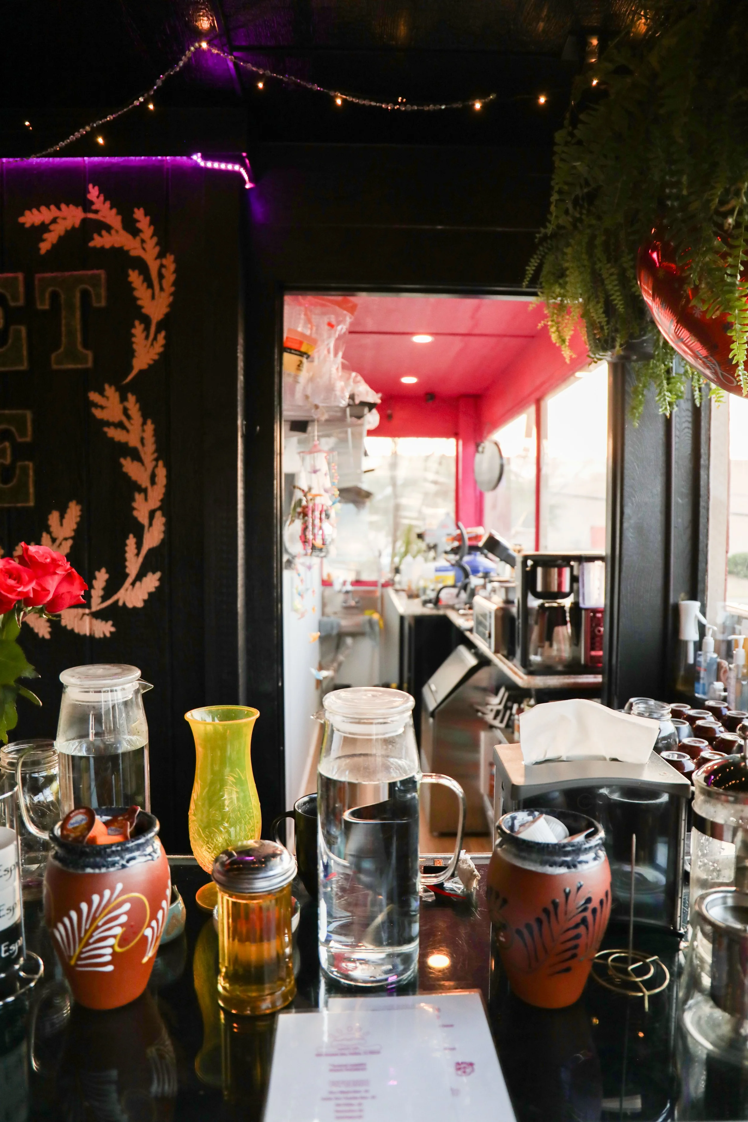 View of a cafe or restaurant interior with various condiments and glasses on the counter. The background shows a kitchen area with equipment, a refrigerator, and a pink ceiling. The decor includes hanging plants and fairy lights.