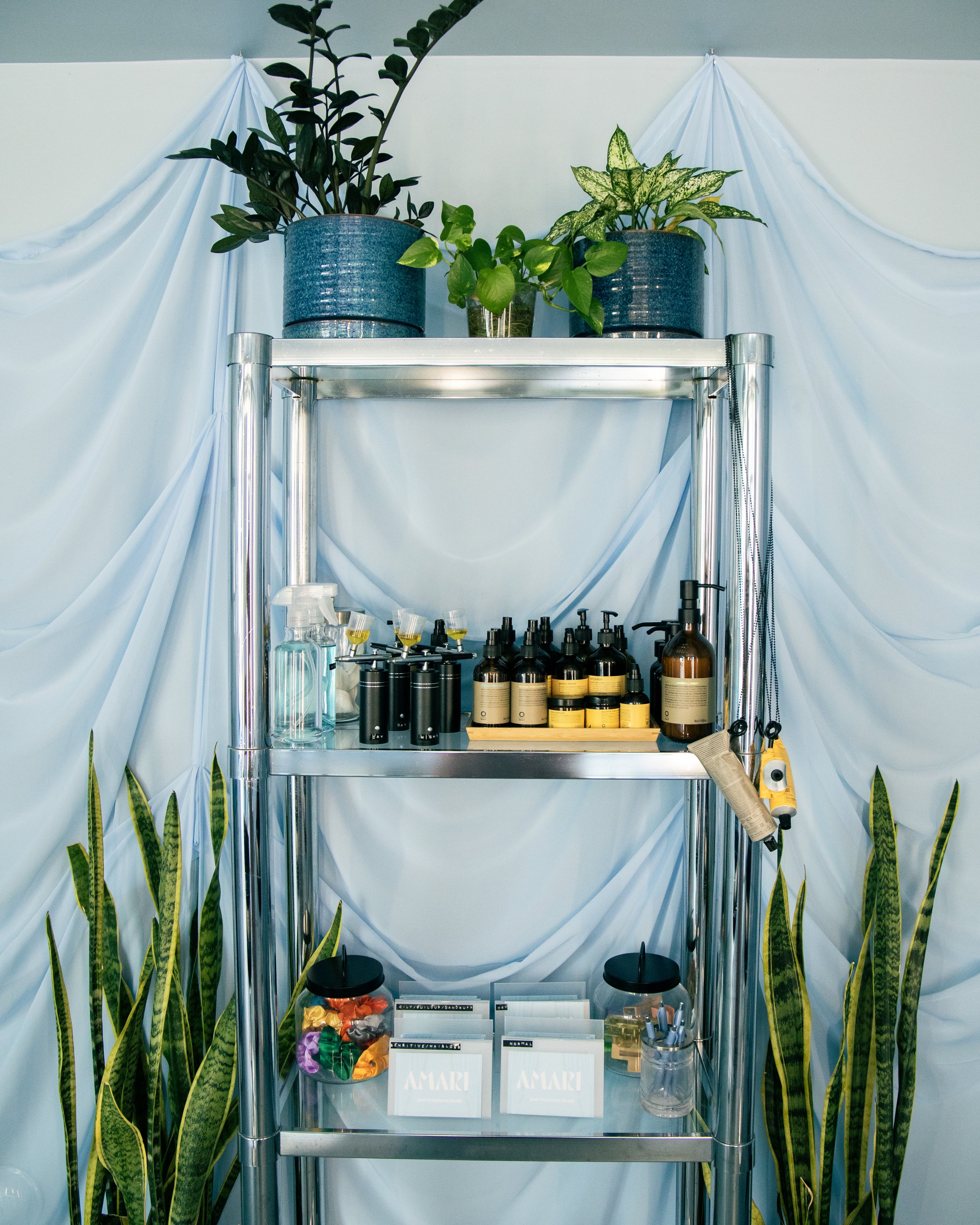A metal shelf with potted plants on top, surrounded by white fabric drapes, and various bottles, jars, and packets on the shelves below.