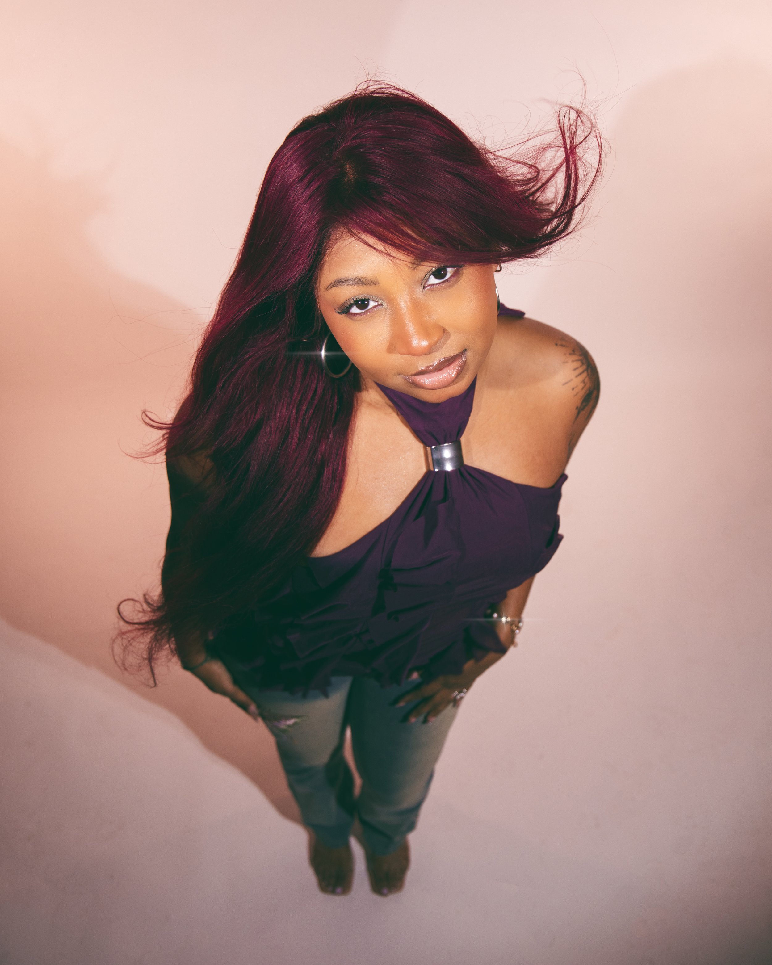 Local Dallas, Texas, DFW Non-traditional Fashion Editorial Creative Portraits Cover Art Photographer Photoshoot. Musician Seihara . A woman with long, dark red hair and hoop earrings, wearing a purple top and jeans, is standing against a light-colore