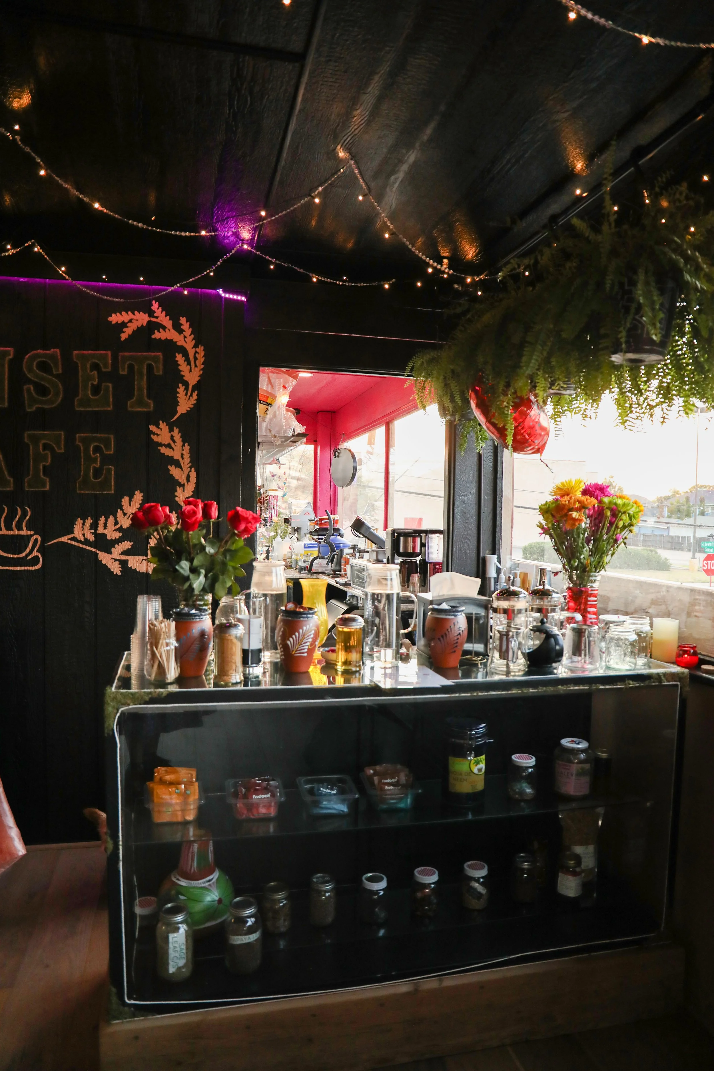 A coffee stand with various syrups, flowers, and jars on display, situated inside a cafe with string lights and plants hanging from the ceiling.