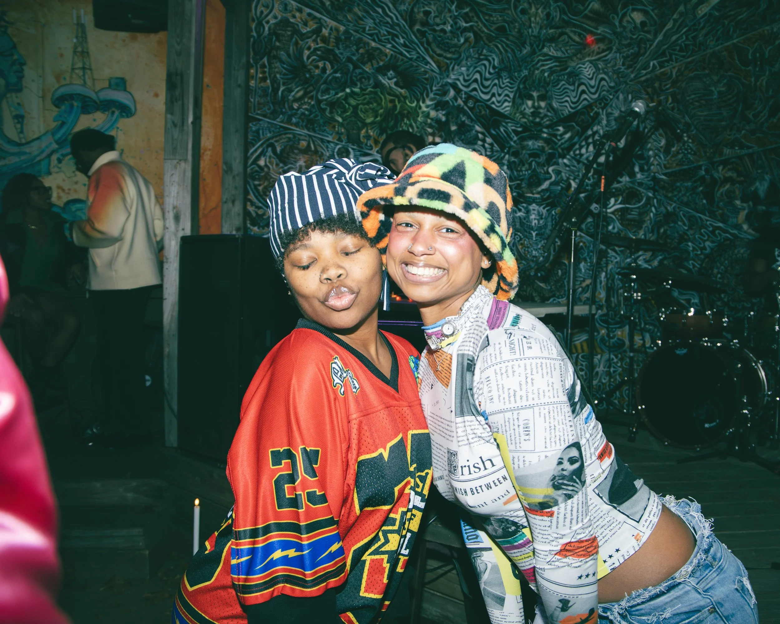 Two young women posing closely together at a lively indoor event, with colorful graffiti art in the background. One woman is wearing a striped beanie and a vibrant, patterned shirt, while the other is wearing a multicolored, animal-print bucket hat a