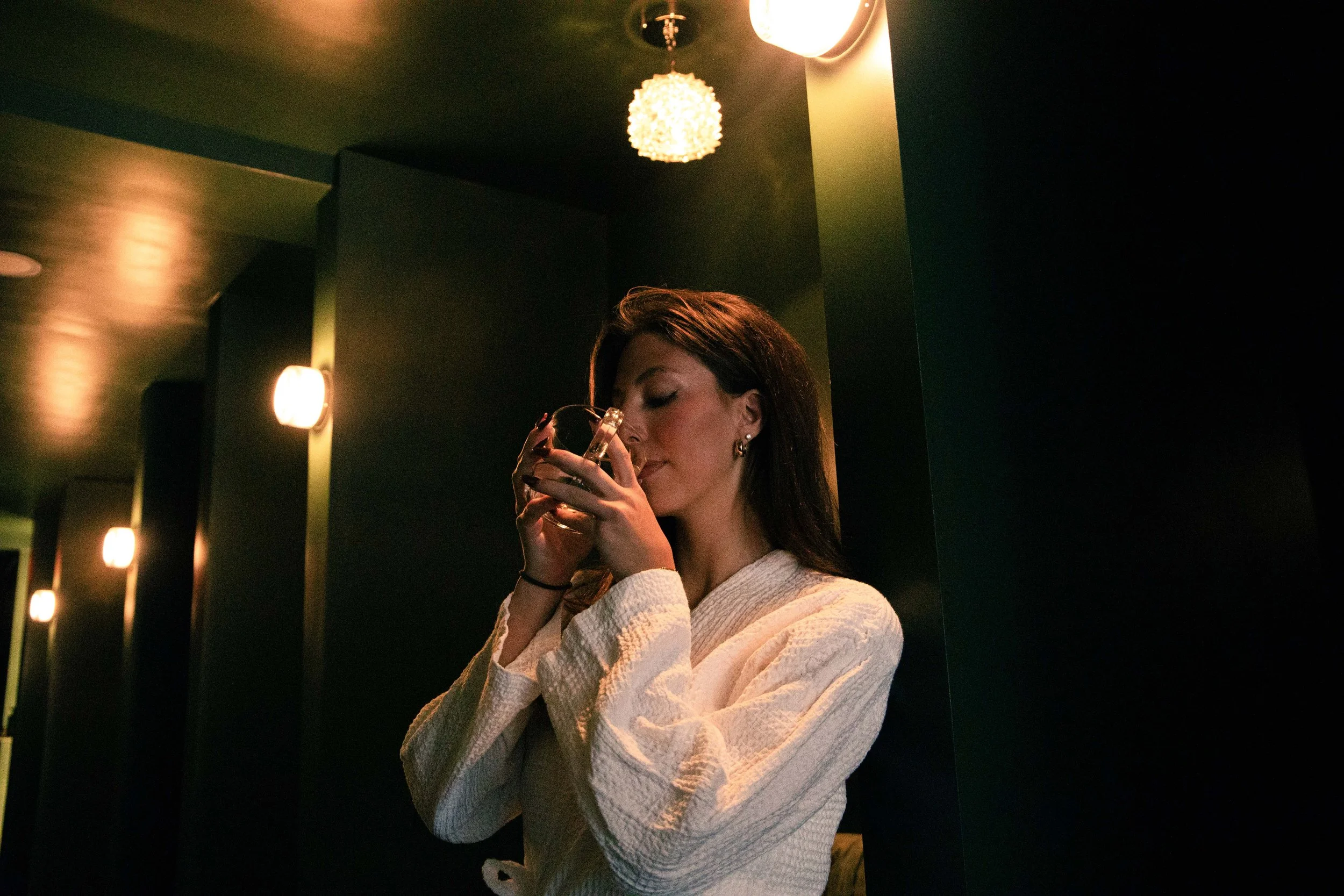 A woman with long brown hair wearing a white sweater, holding a glass of drink, standing in a dimly lit room with warm lighting and modern decor, including hanging light fixtures.