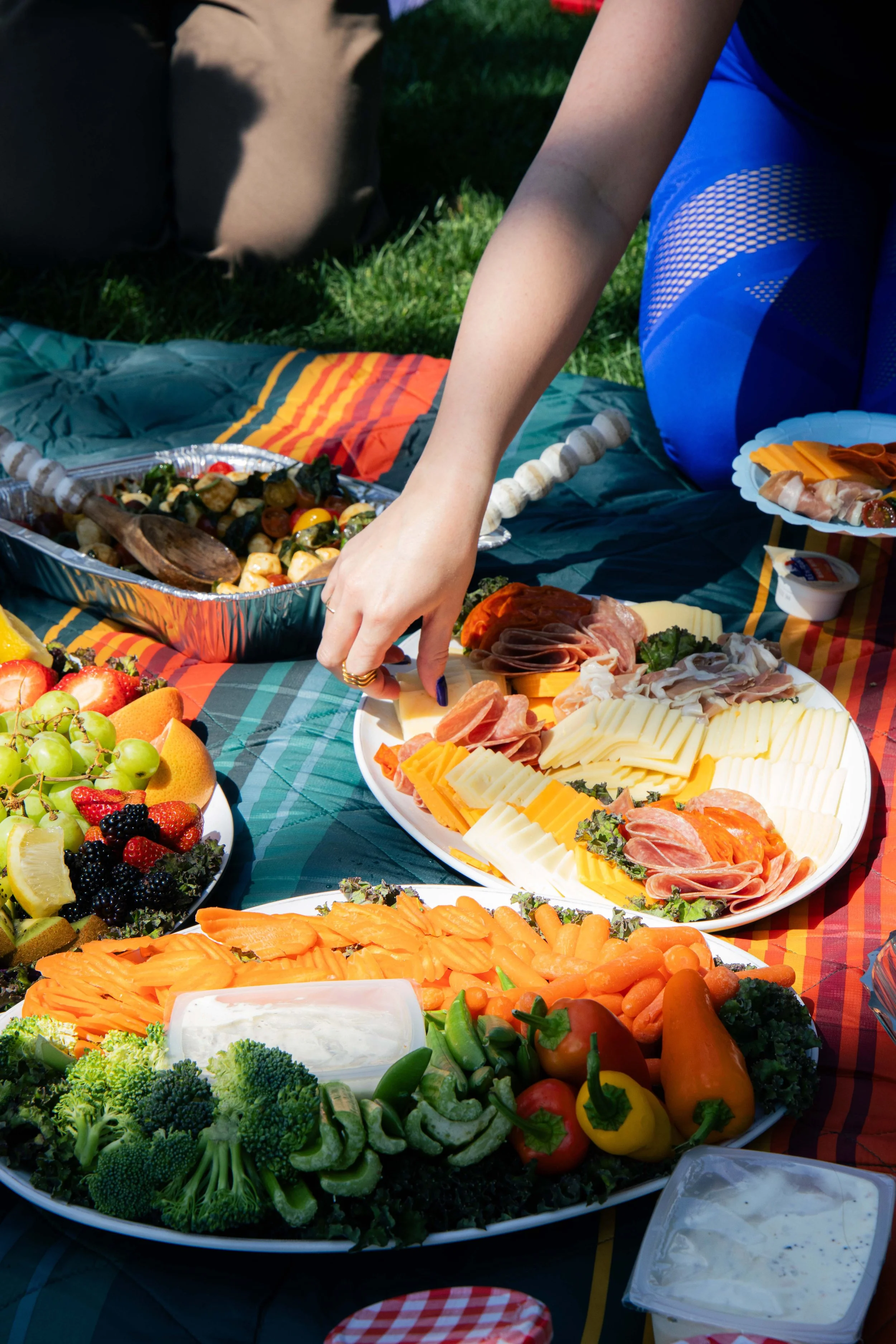 A picnic spread on a grassy lawn with various dishes, including a vegetable platter with broccoli, carrots, and yellow peppers, a cheese and meat platter, and fruit bowls, with someone reaching for food.