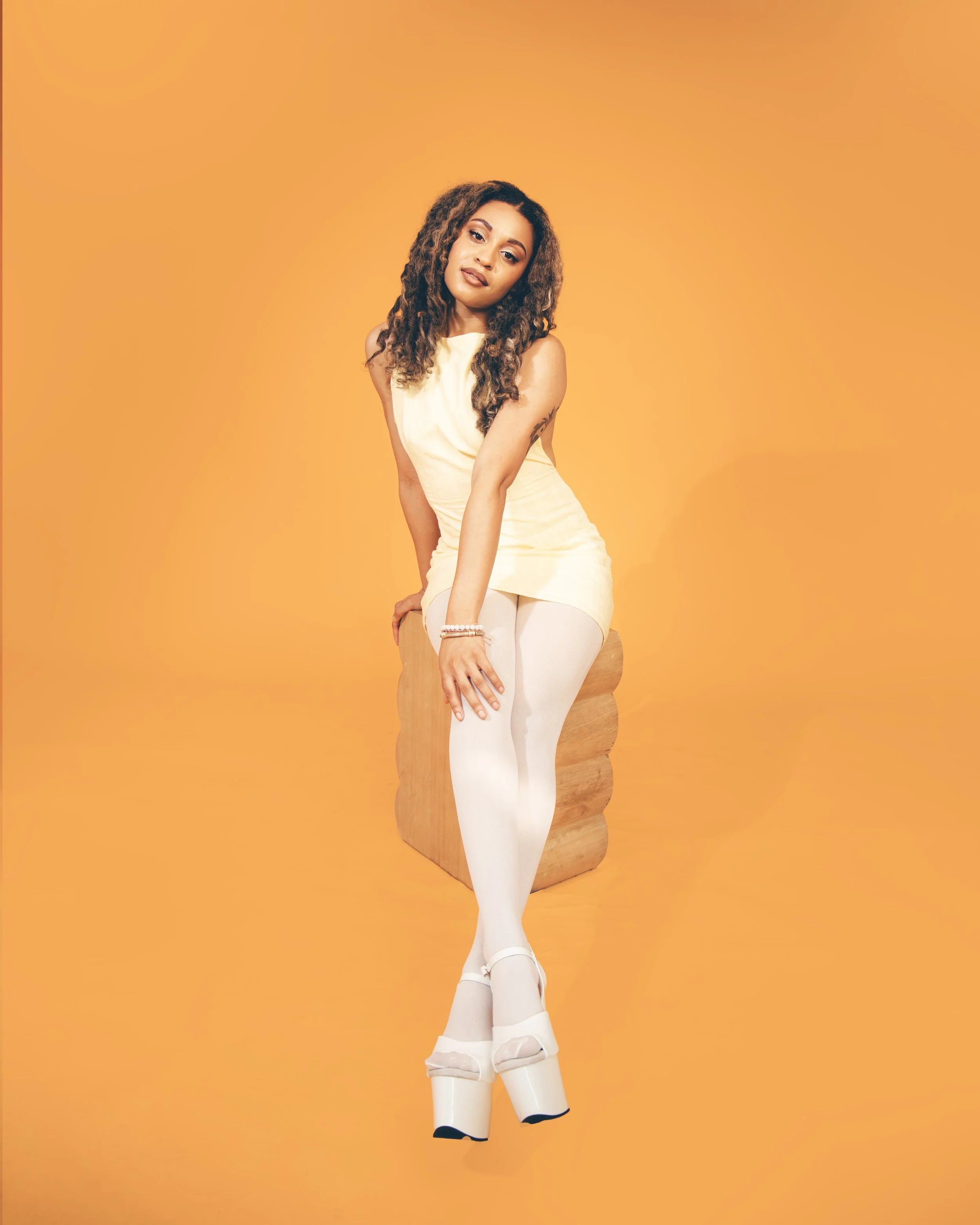 Local Dallas, Texas, DFW Non-traditional Fashion Editorial Creative Portraits Cover Art Photographer Photoshoot. Musician Rebelle Flor . A woman with brown curly hair wearing a cream sleeveless dress and white tights, sitting on a wooden chair agains