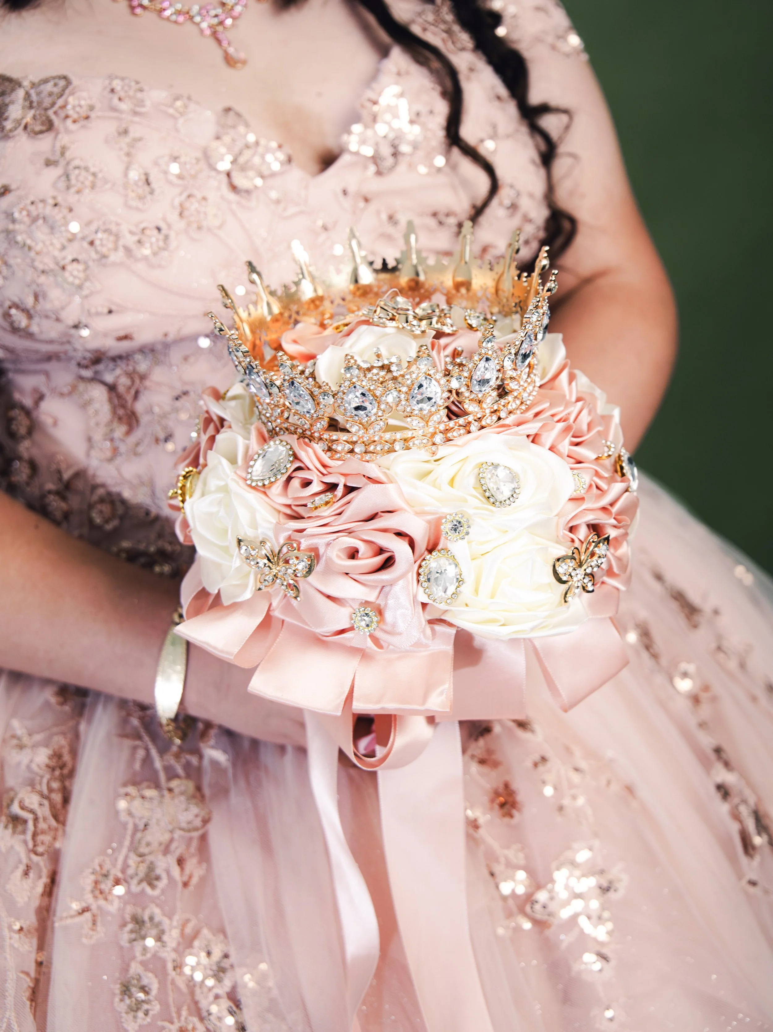 Close-up of a young girl in a pink floral dress holding a bouquet with a tiara. The bouquet is decorated with rhinestones, pearls, and butterfly ornaments, wrapped in pink ribbon ribbons.