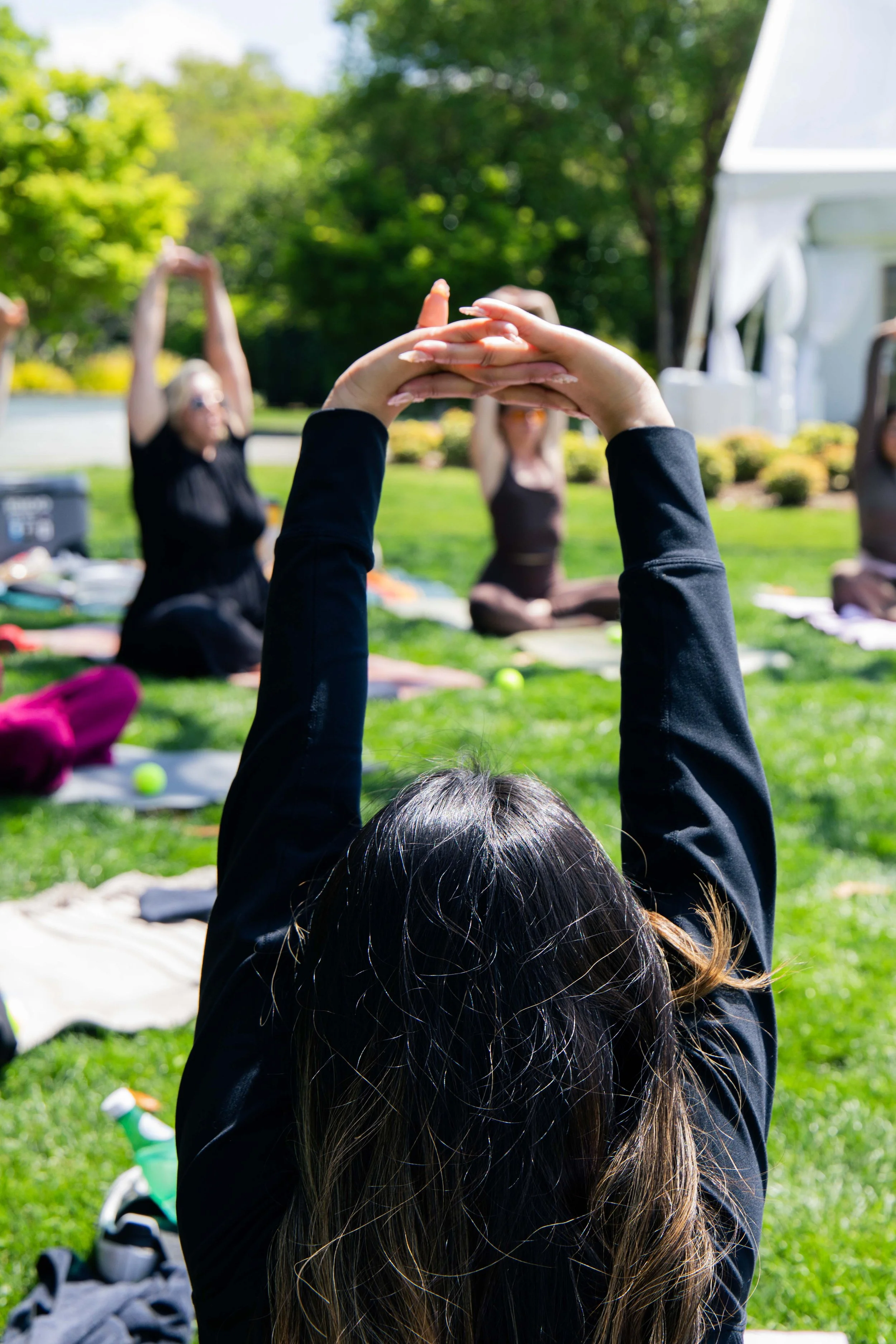 People practicing yoga outdoors on a sunny day, sitting on mats in a park with green trees in the background.