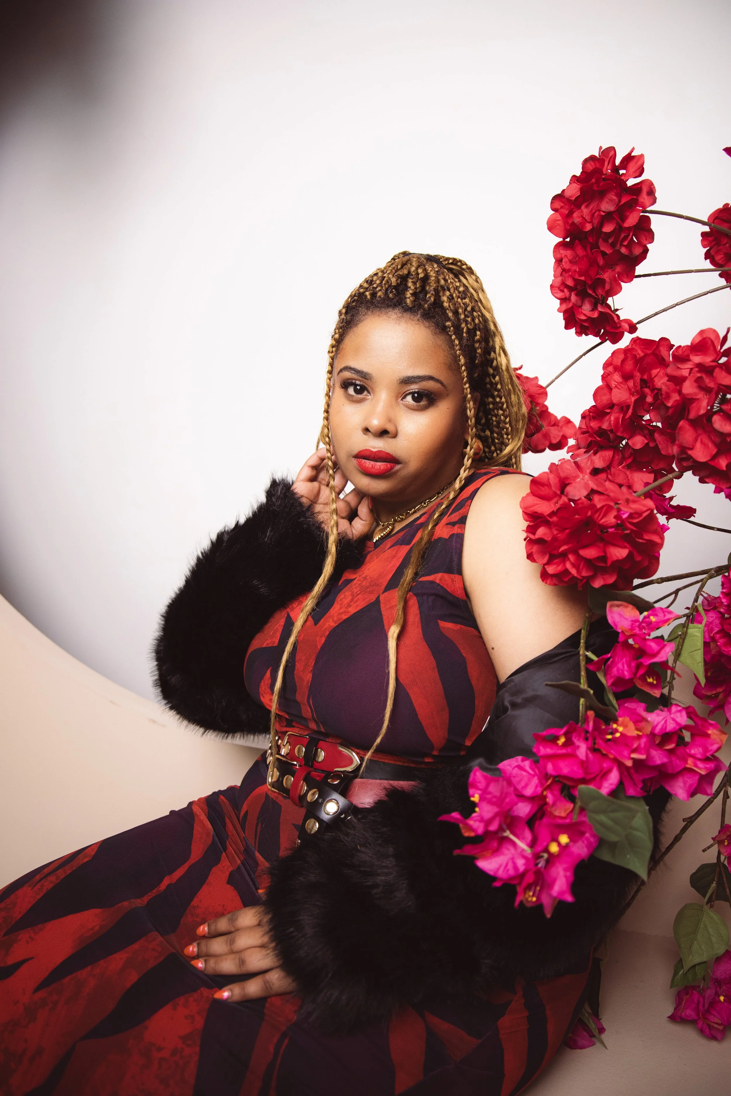Local Dallas, Texas, DFW Non-traditional Fashion Editorial Creative Portraits Cover Art Photographer Photoshoot. A woman with braided blonde hair, red lipstick, and a black and red outfit, sitting beside pink flowers against a plain background.