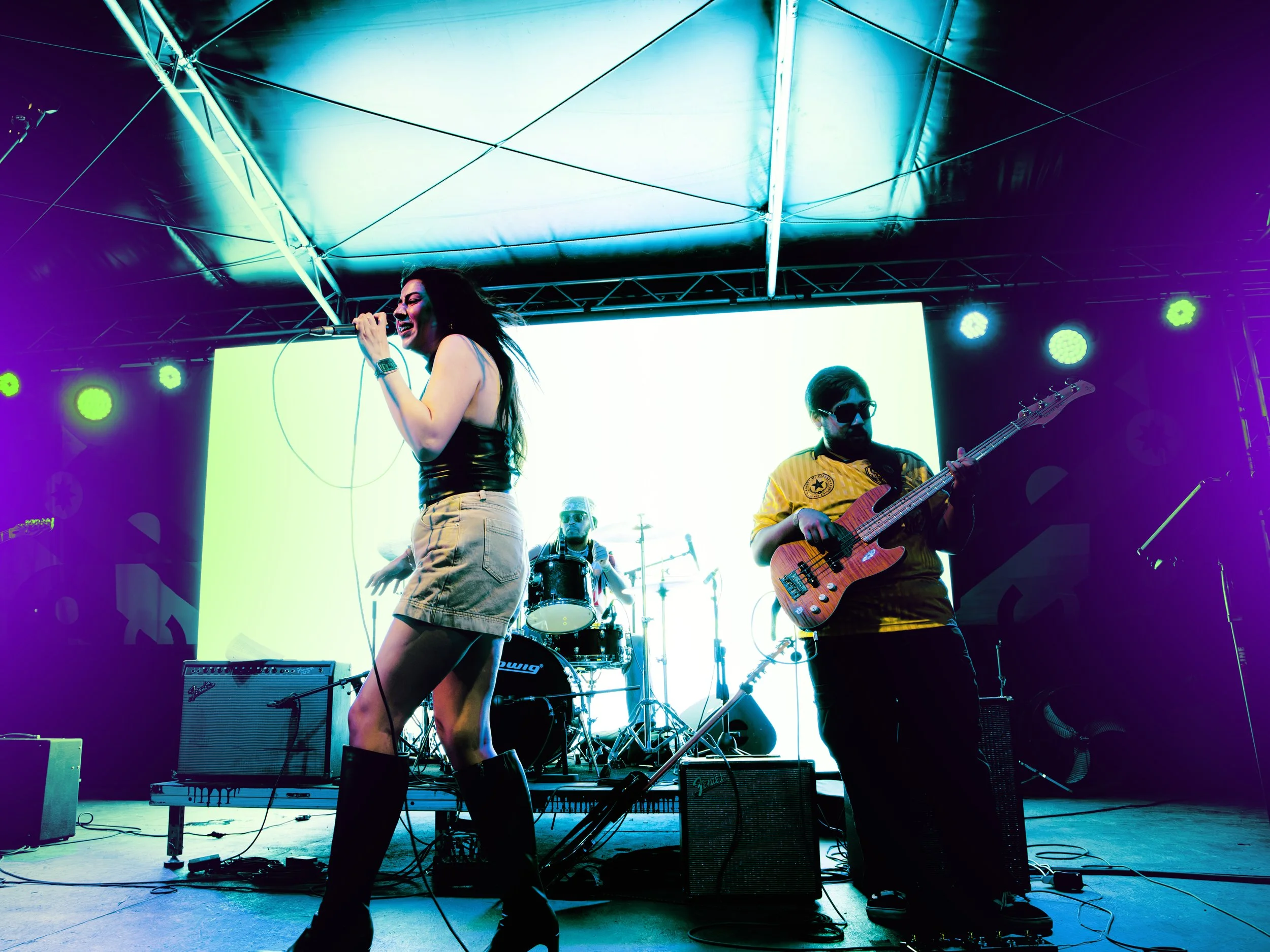 Musicians performing on a brightly lit stage with colorful lights. A female singer with long hair, black outfit, and boots sings into a microphone. A male guitarist wearing sunglasses and a yellow shirt plays bass guitar. A drummer can be seen in the