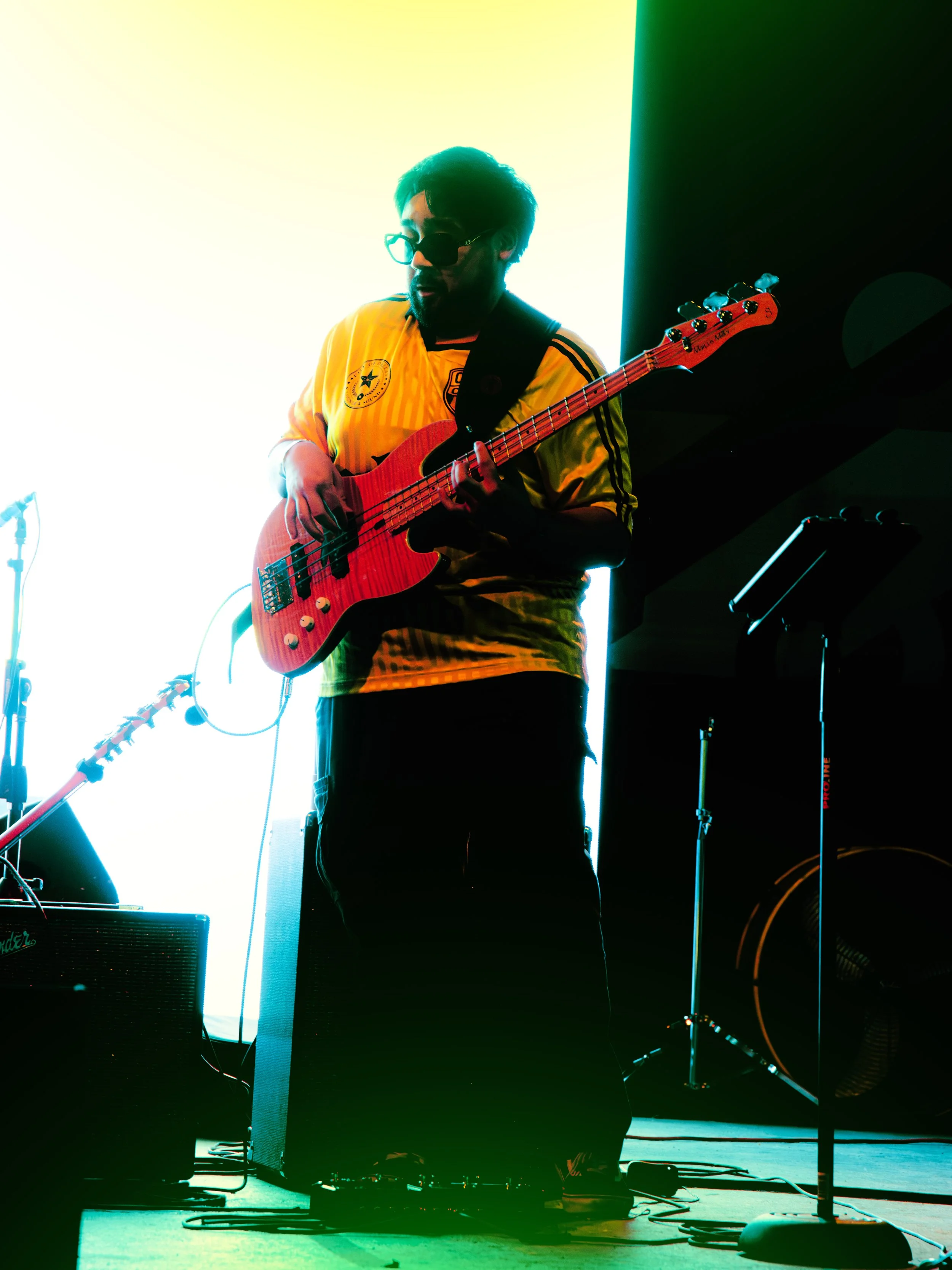 A person playing an electric bass guitar on stage, wearing sunglasses and a yellow sports jersey, with stage lights creating a colorful backdrop.
