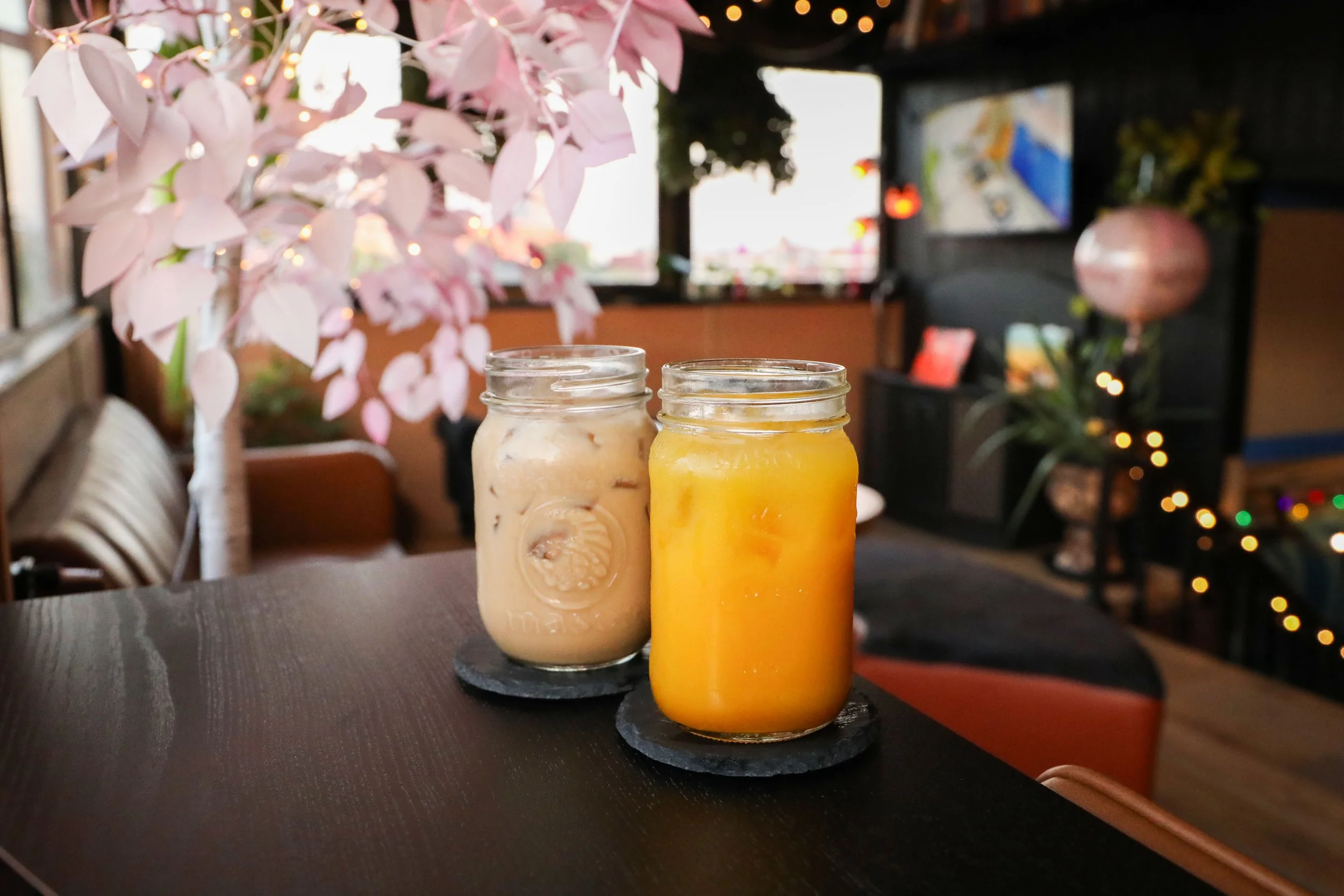 Three mason jars filled with creamy beverages and a bright orange juice, placed on black round coasters on a dark wood table in a cozy indoor setting with pink flowers, warm lighting, and a television in the background.