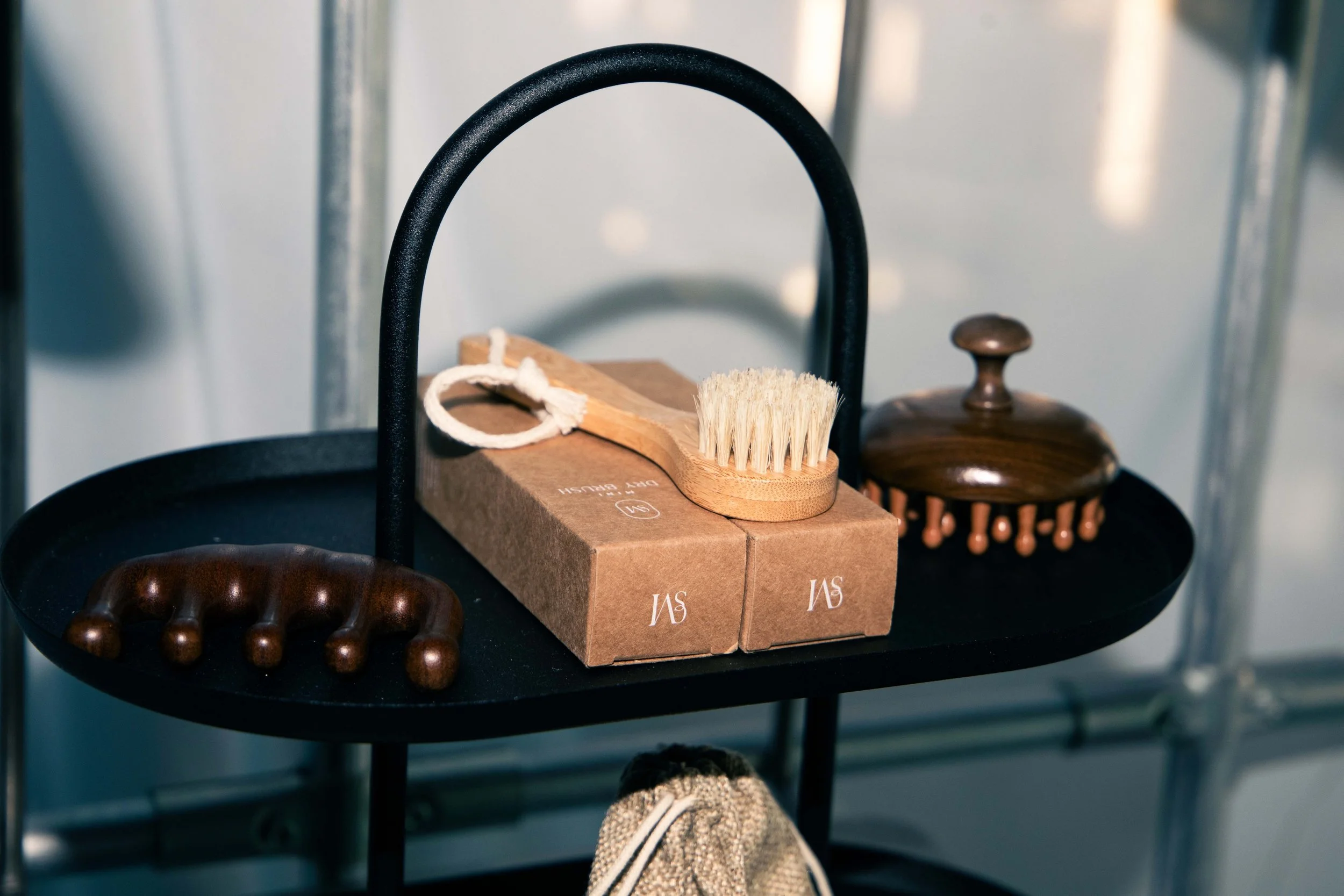 Close-up of a shoe on a person's foot on a black circular table, with a wooden brush and two cardboard boxes with white text, behind the table, with a metal background and a blurred wooden handrail or part of a furniture piece.