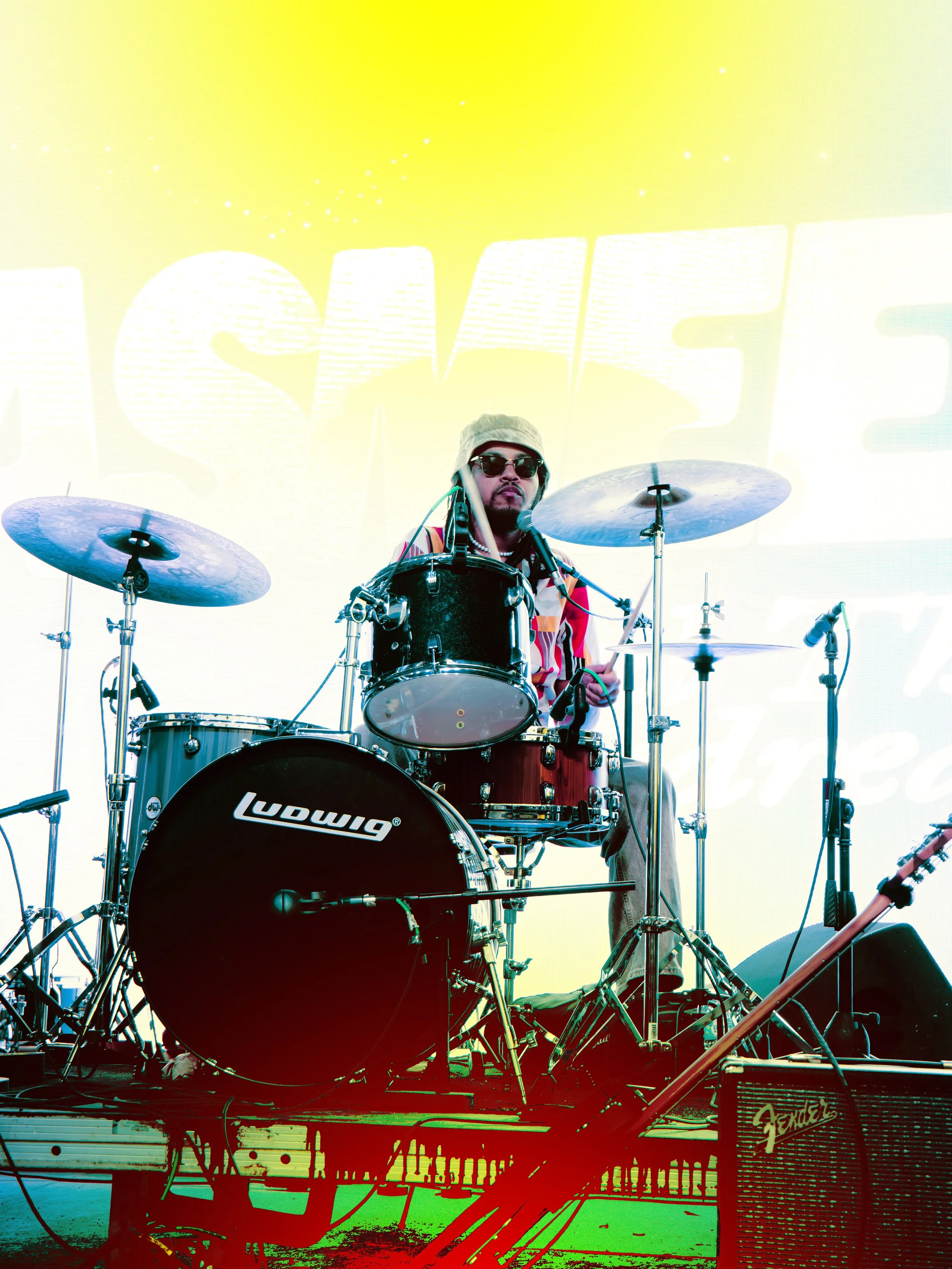 A musician wearing sunglasses, a bucket hat, and a striped shirt playing a drum set on a stage with a bright yellow background and large partial text behind him.