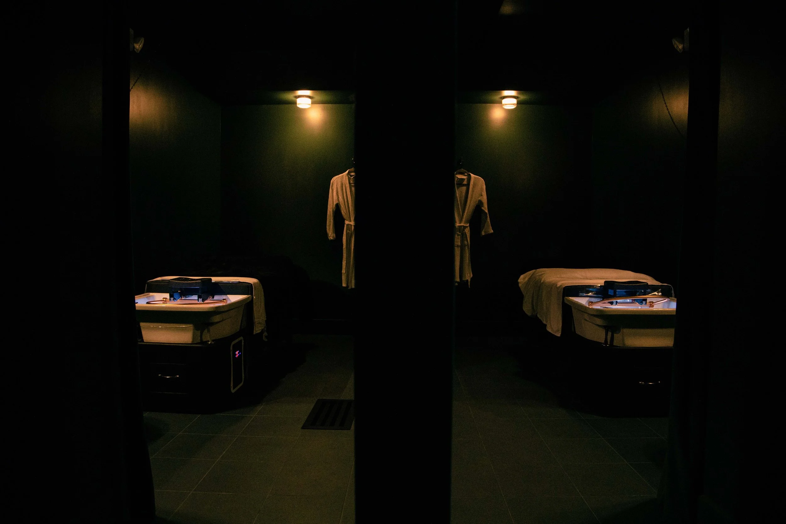 Dark locker room with two bathrobes hanging on the wall and two sinks on either side, and a bathtub in the foreground.