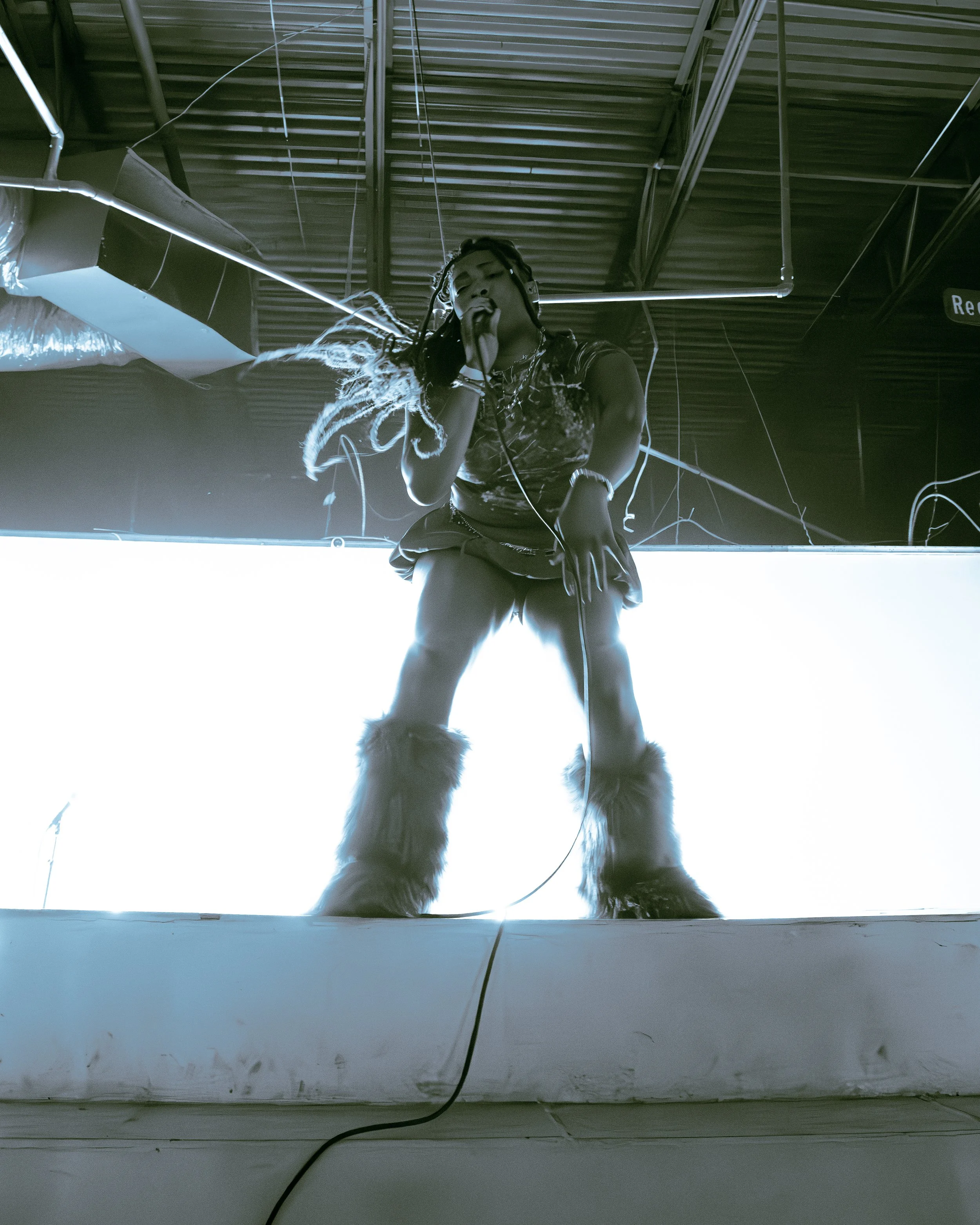 A person wearing furry boots crouching down, holding a microphone, and performing on a stage with a reflective surface. The background shows a ceiling with exposed pipes and lights.
