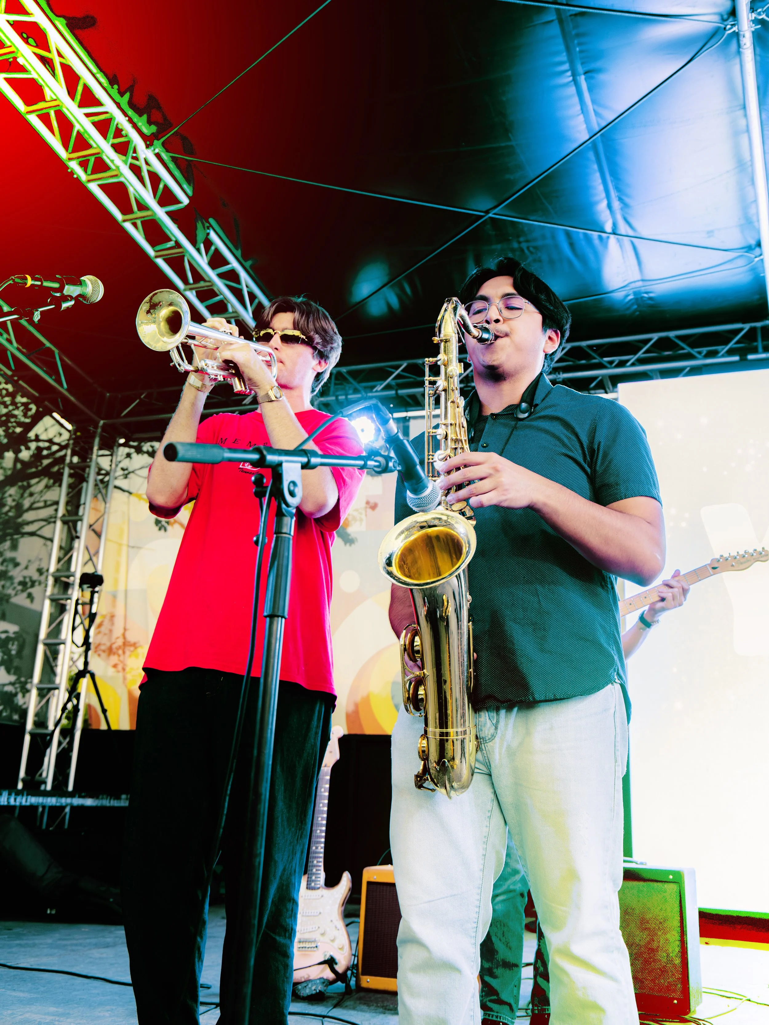 Two musicians playing instruments on stage: one with a trumpet wearing sunglasses and a red shirt, and the other with a saxophone wearing glasses and a dark shirt, under a black tent with stage lighting and musical equipment.
