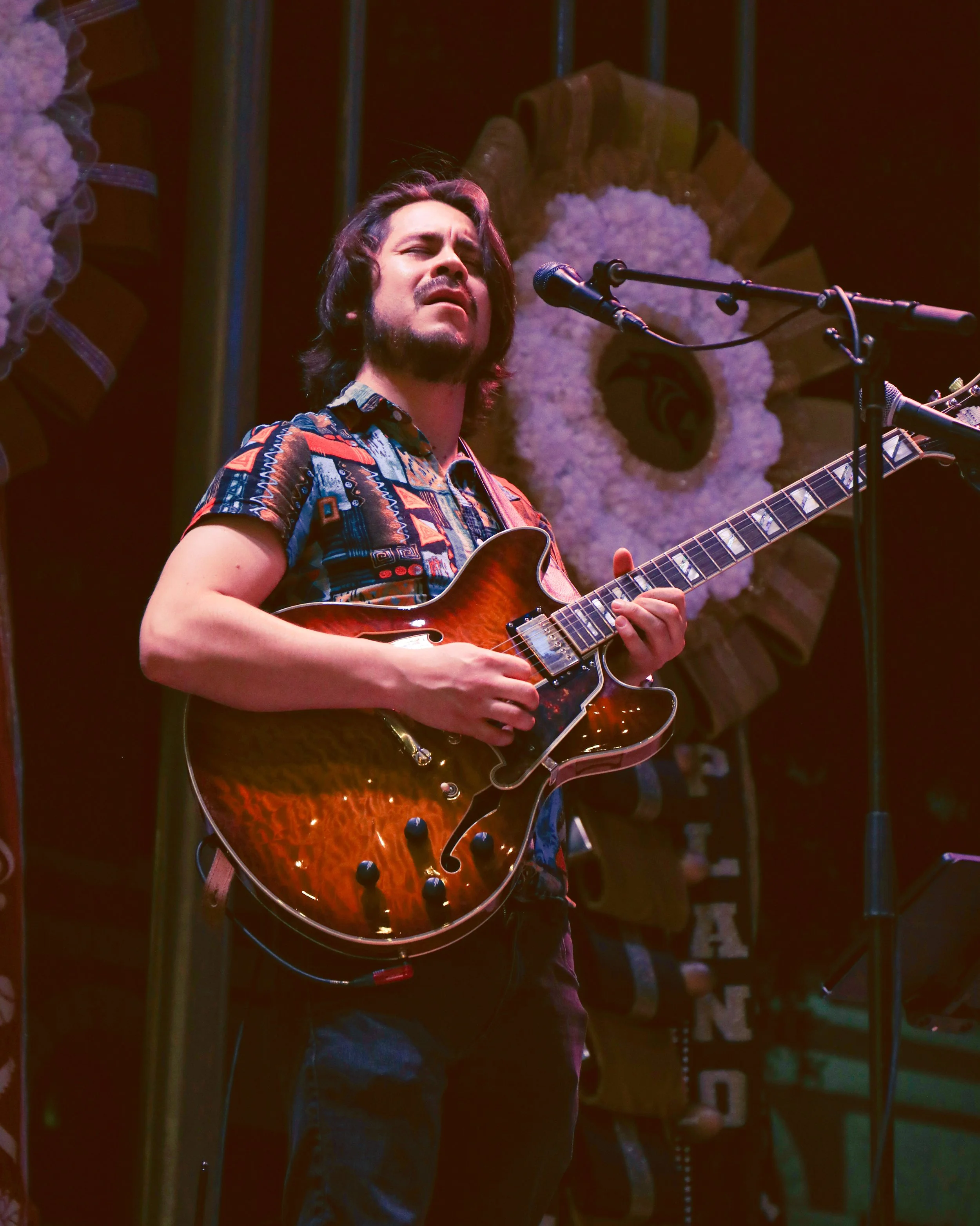 A man with shoulder-length dark hair and a beard, wearing a colorful patterned shirt, is singing and playing an electric guitar on stage. Behind him is a decorative floral arrangement with white flowers and a crown-shaped ornament.