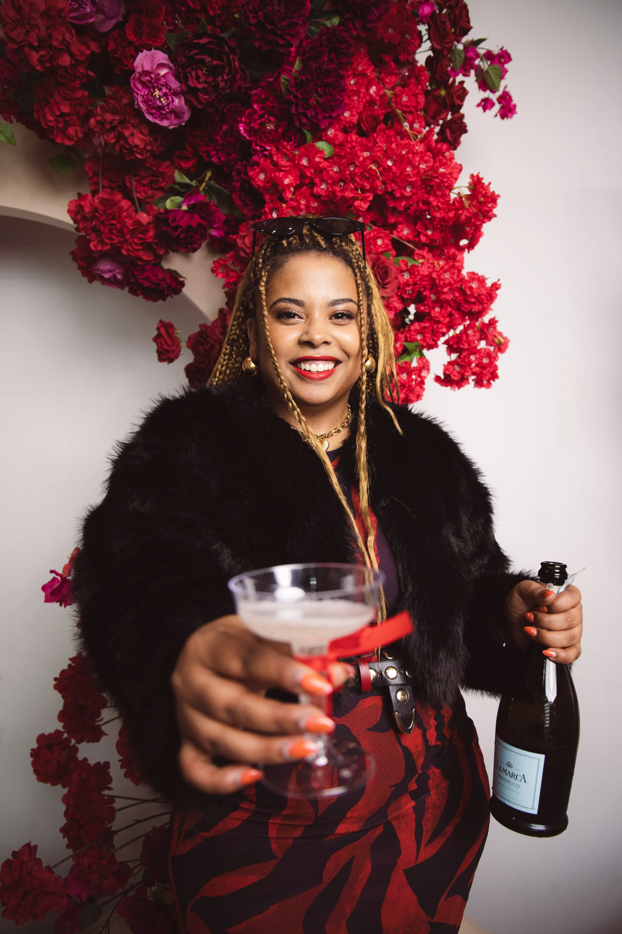 A woman with blonde braided hair smiling, wearing a black faux fur coat, holding a martini glass and a bottle of sparkling wine, in front of a vibrant red and pink floral backdrop.