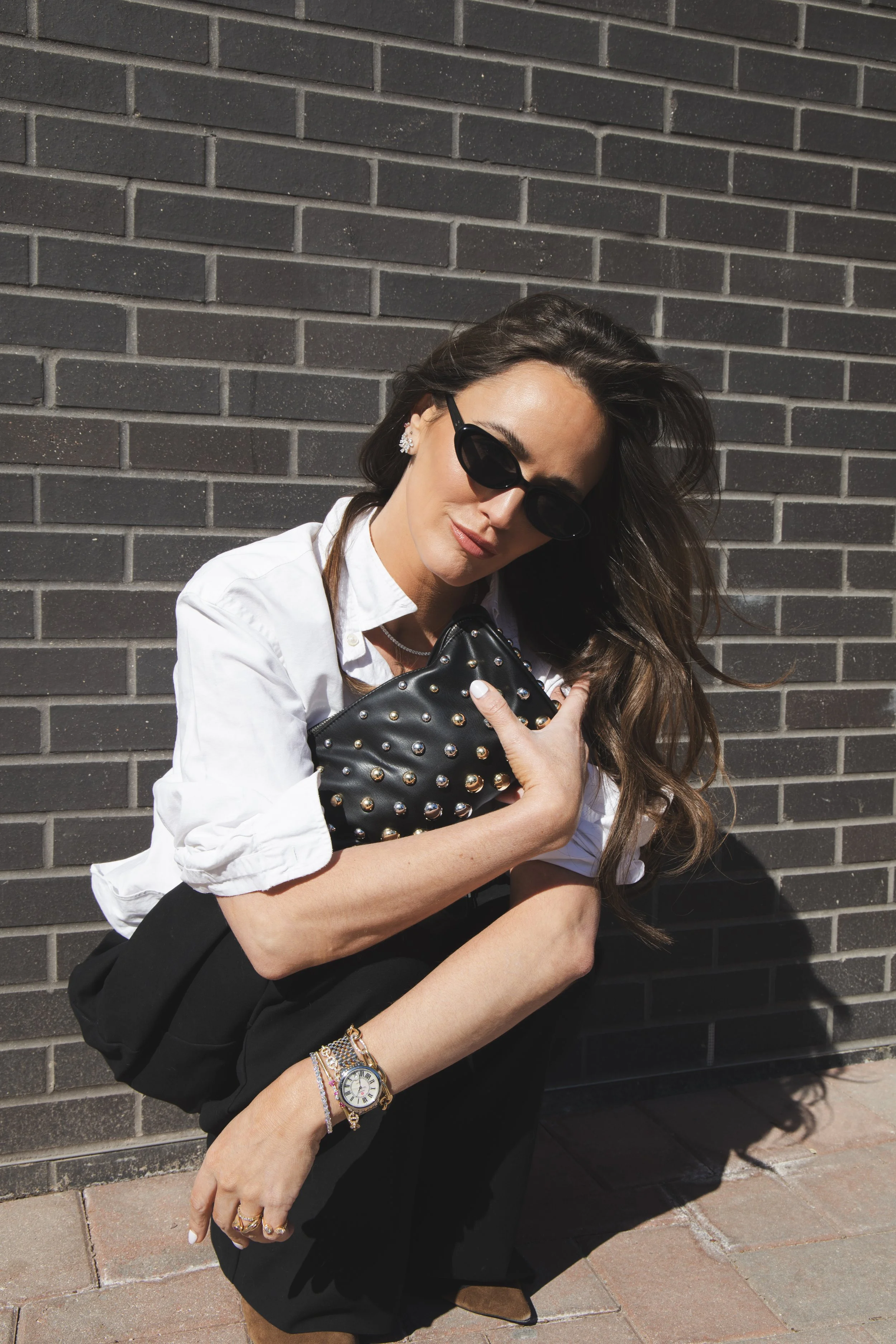 Local Dallas, Texas, DFW Non-traditional Fashion Editorial Creative Portraits Cover Art Photographer Photoshoot. Revel Workshop . A woman with long, wavy hair wearing dark sunglasses, a white shirt, and black pants, holding a studded black purse, sta