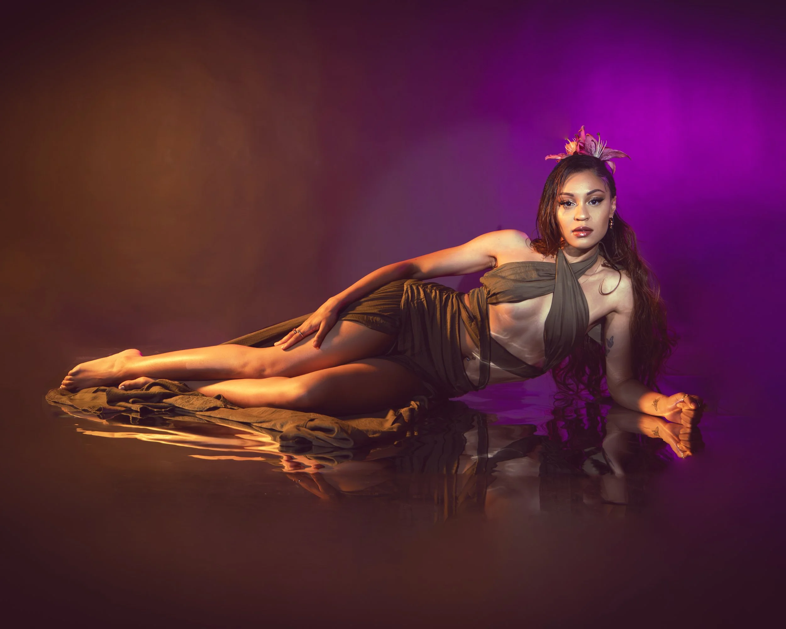 Local Dallas, Texas, DFW Non-traditional Editorial Creative Portraits Cover Art Photographer. musician Rebelle Flor . A woman with long dark hair and makeup, wearing a dress, lying on a reflective surface with purple and orange lighting effect, and a