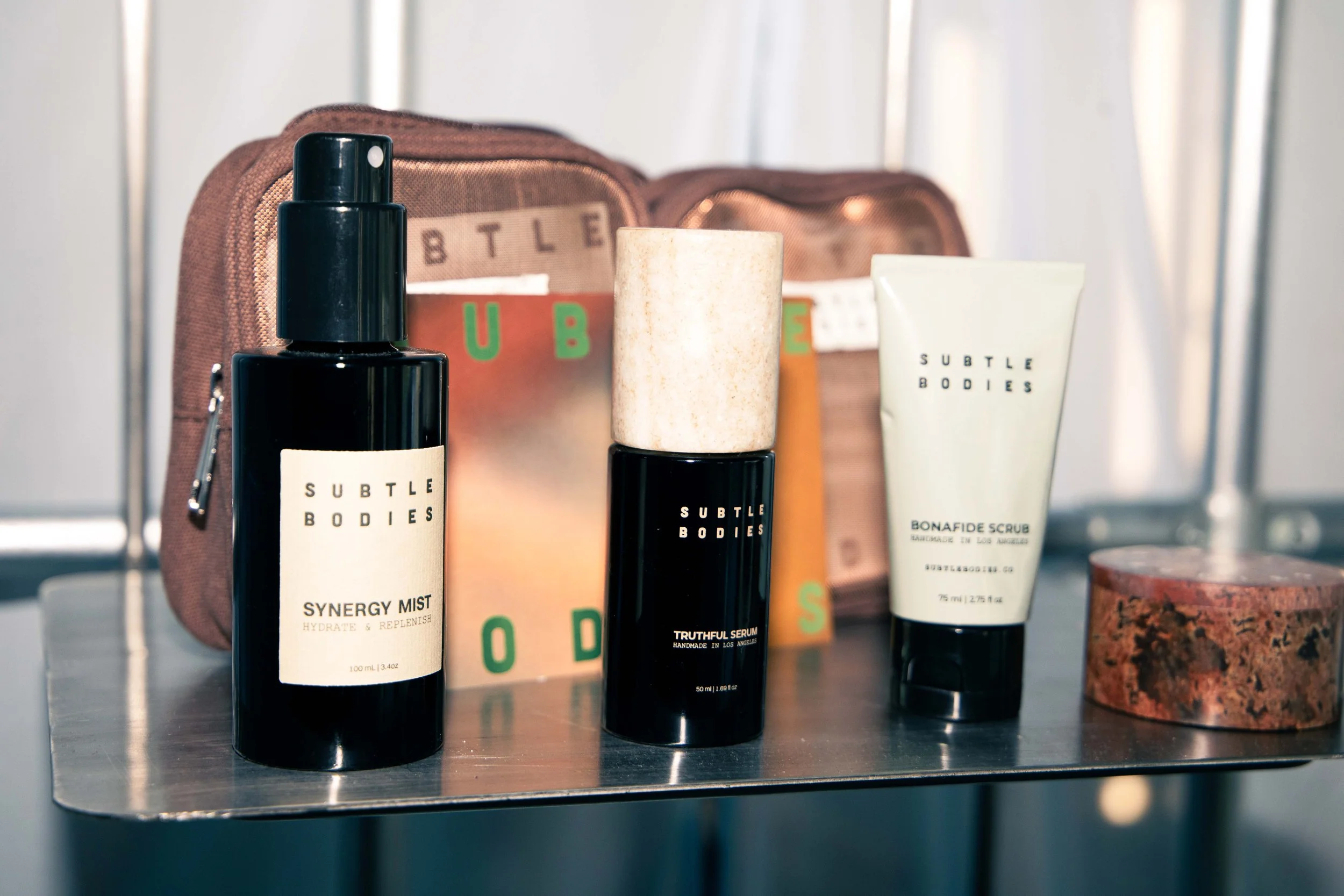 A collection of skincare products on a metallic surface, including bottles labeled 'Subtle Bodies' and a pouch in the background.