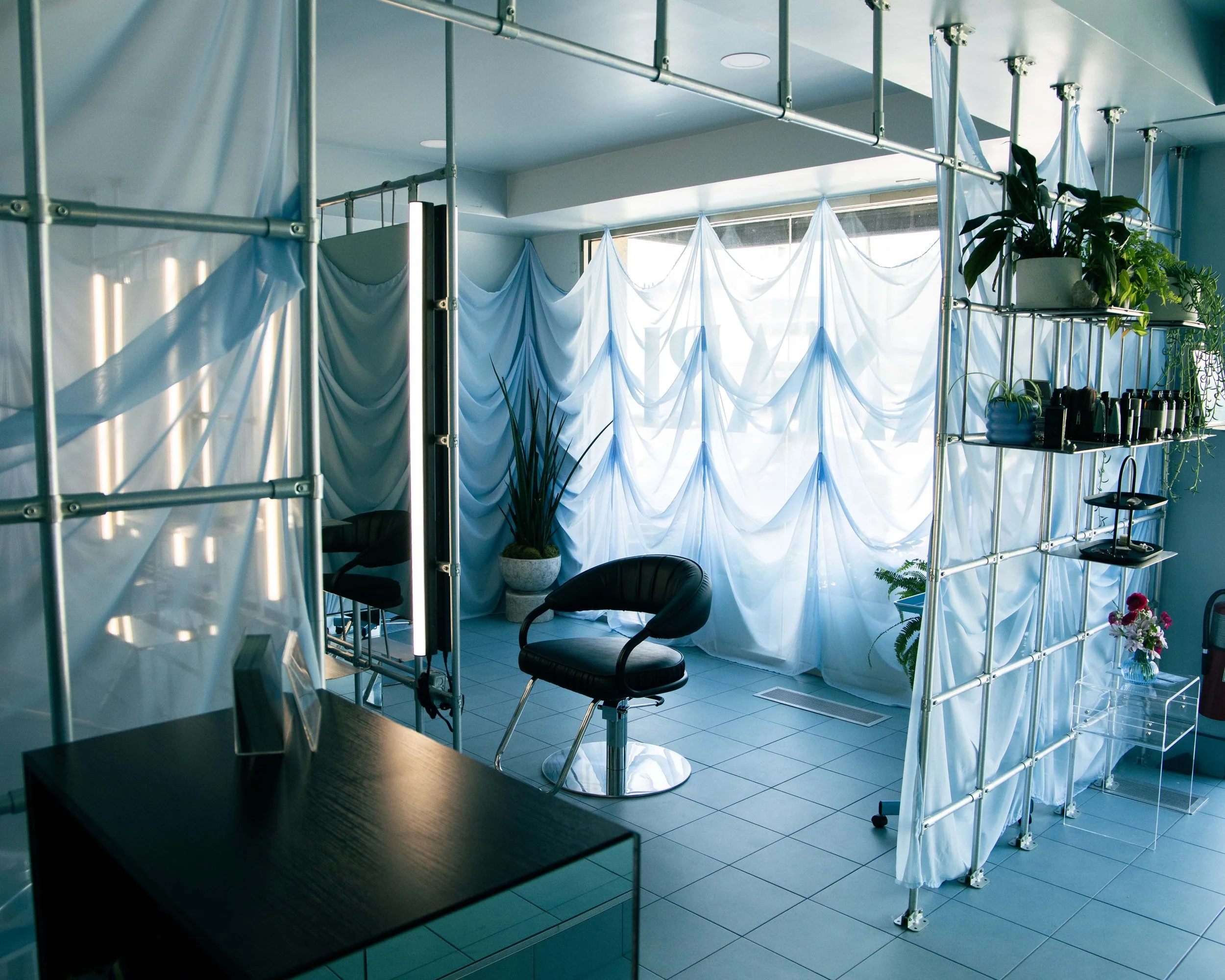 Interior of a modern space with a metal scaffold supporting white and blue draped curtains, sunlight coming through large windows, and various potted plants on shelves and the floor.