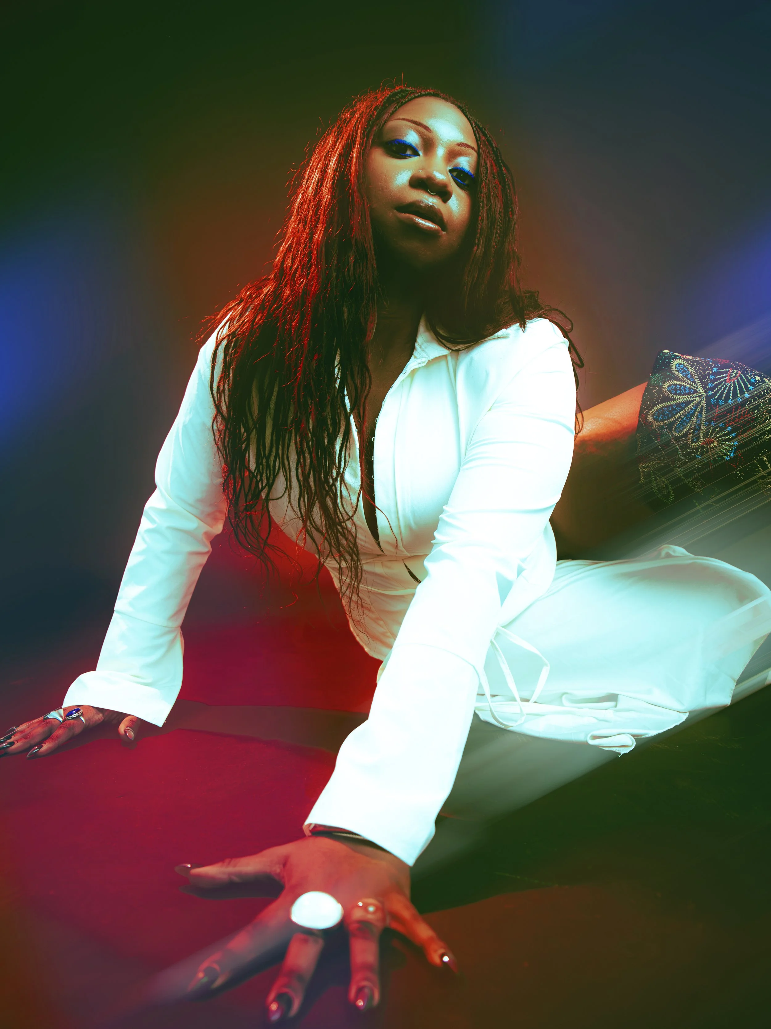 Local Dallas, Texas, DFW Non-traditional Fashion Editorial Creative Portraits Cover Art Photographer Photoshoot. Musician Courtney noir. A woman with long dreadlocks and blue eyeshadow, sitting on a surface, with a hand extended towards the camera, w