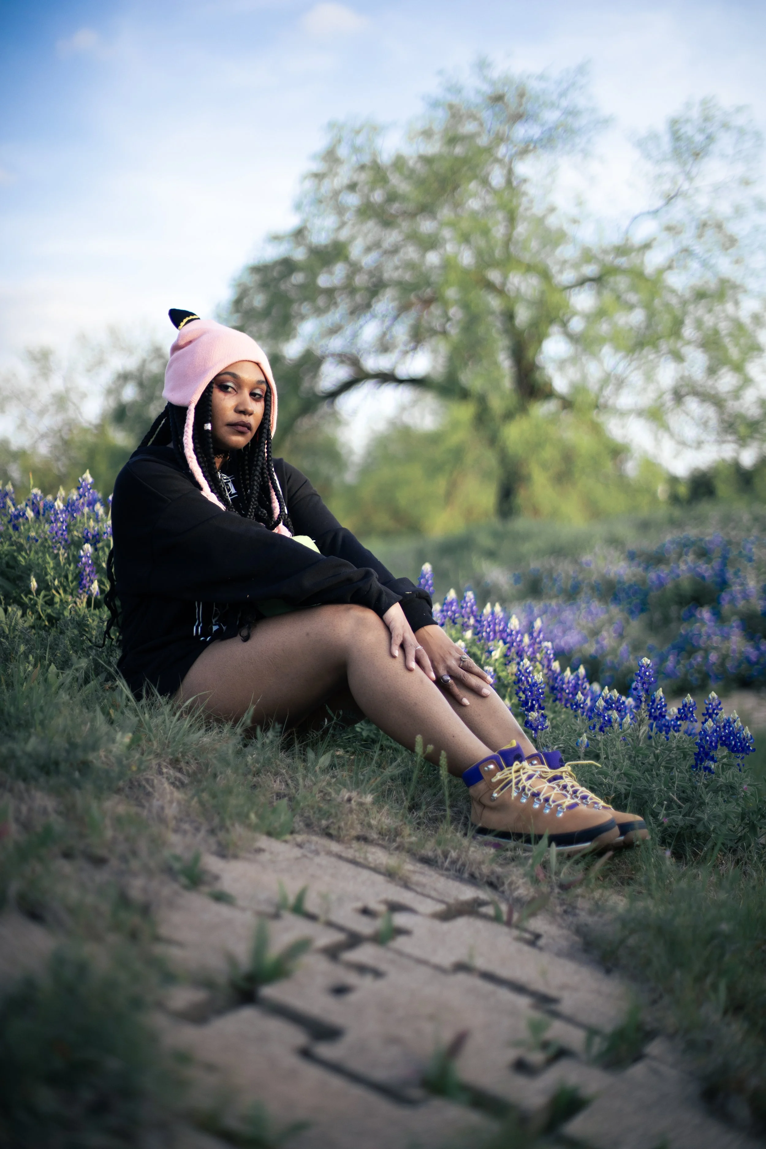 Local Dallas, Texas, DFW Non-traditional Fashion Editorial Creative Portraits Cover Art Photographer Photoshoot. musician igimeji . A young woman with dark skin, braided hair, and a pink beanie hat with earflaps, sitting on the grass among purple flo