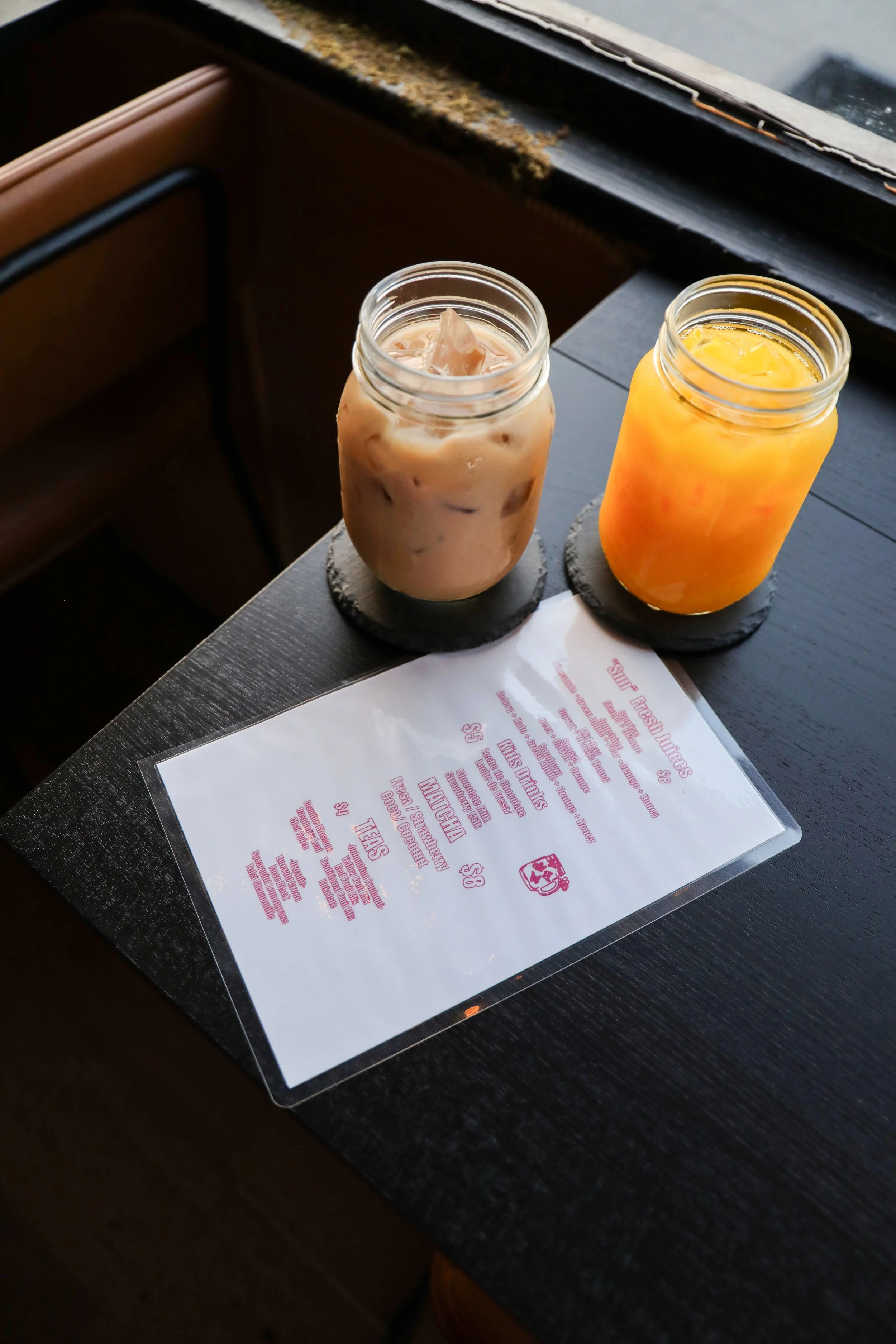 Two mason jars filled with iced tea or juice on a dark table, next to a menu with red text, near a window with a black frame.